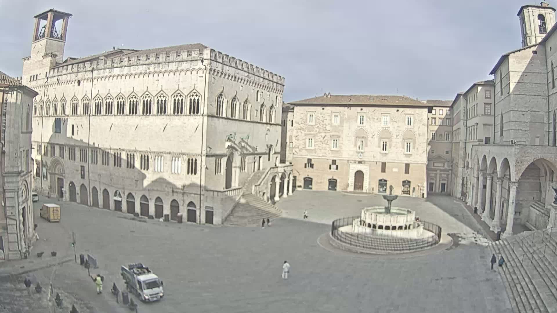 A large, mostly empty town square is surrounded by several aged stone buildings, one of which features a prominent staircase, under a bright, clear sky.