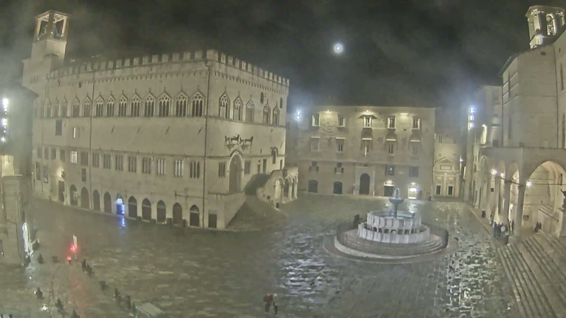 A large, stone plaza at night, wet from rain, features a central fountain and is surrounded by historic buildings.