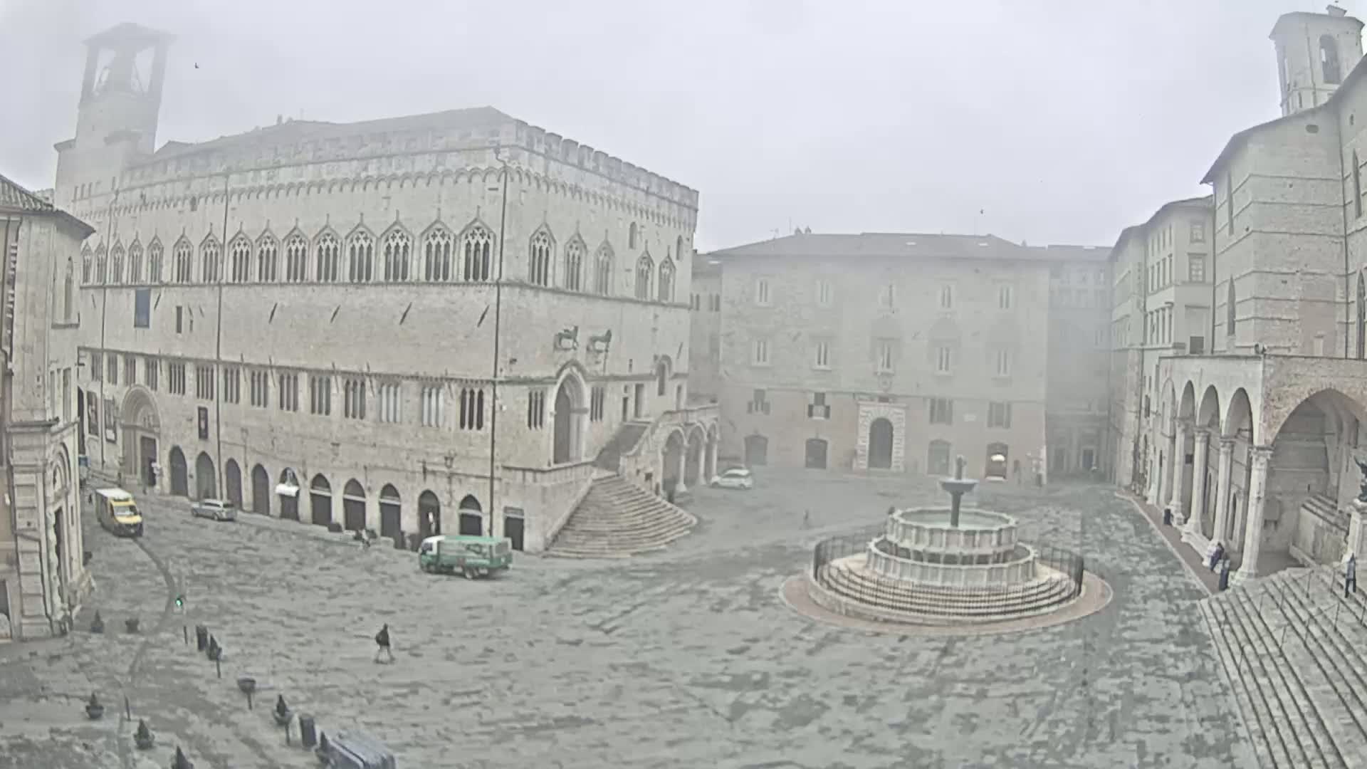 A large, stone plaza with a fountain is shrouded in fog, featuring several historic buildings surrounding it.