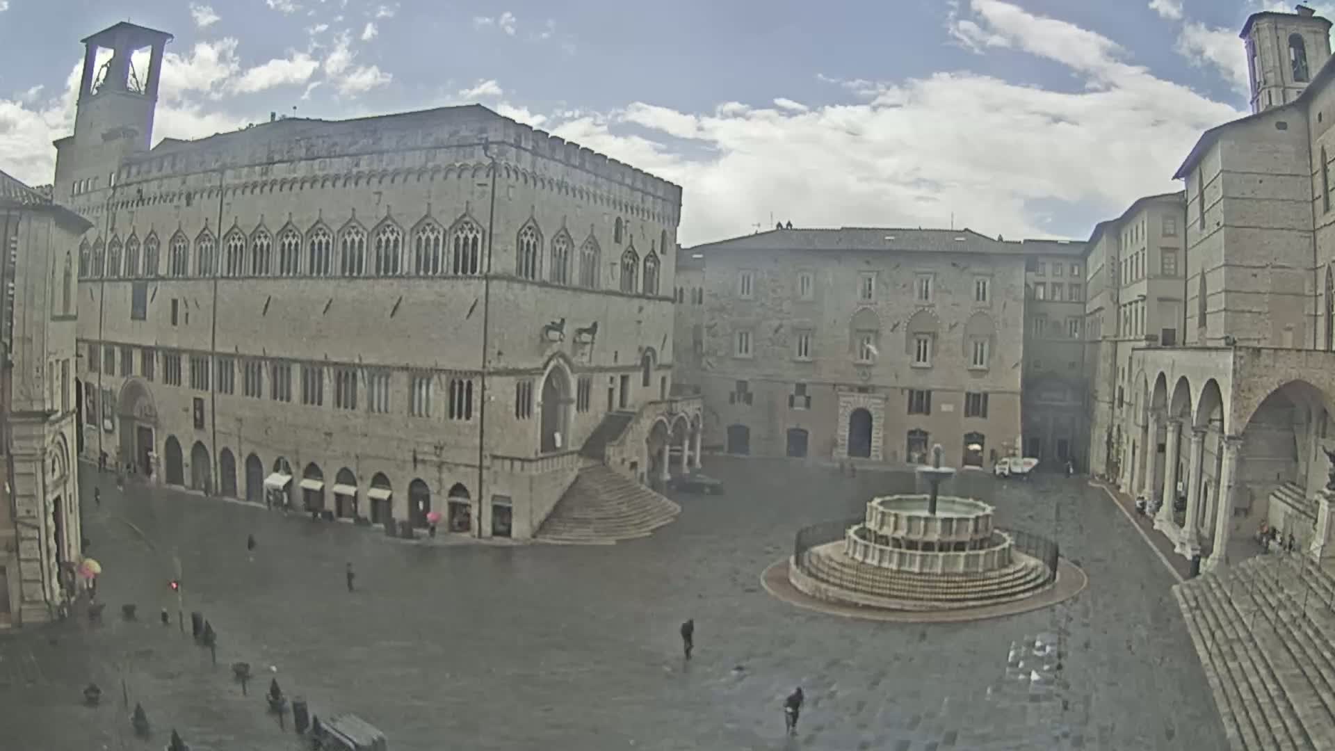 A large, stone square with a fountain in the center, surrounded by medieval-style buildings under a partly cloudy sky.