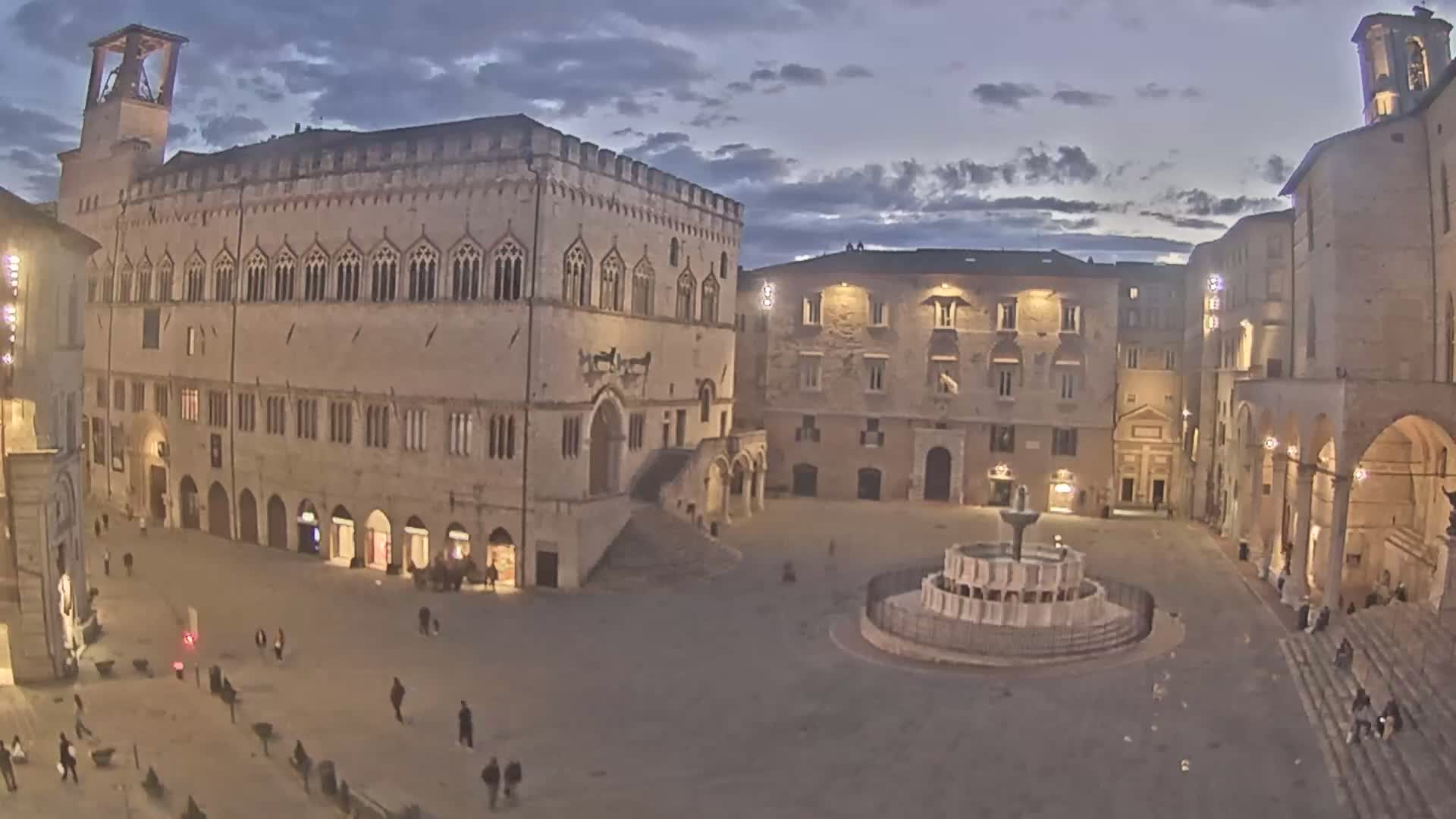 A large, stone plaza at twilight, featuring a central fountain and several illuminated buildings, is mostly empty except for a few pedestrians.
