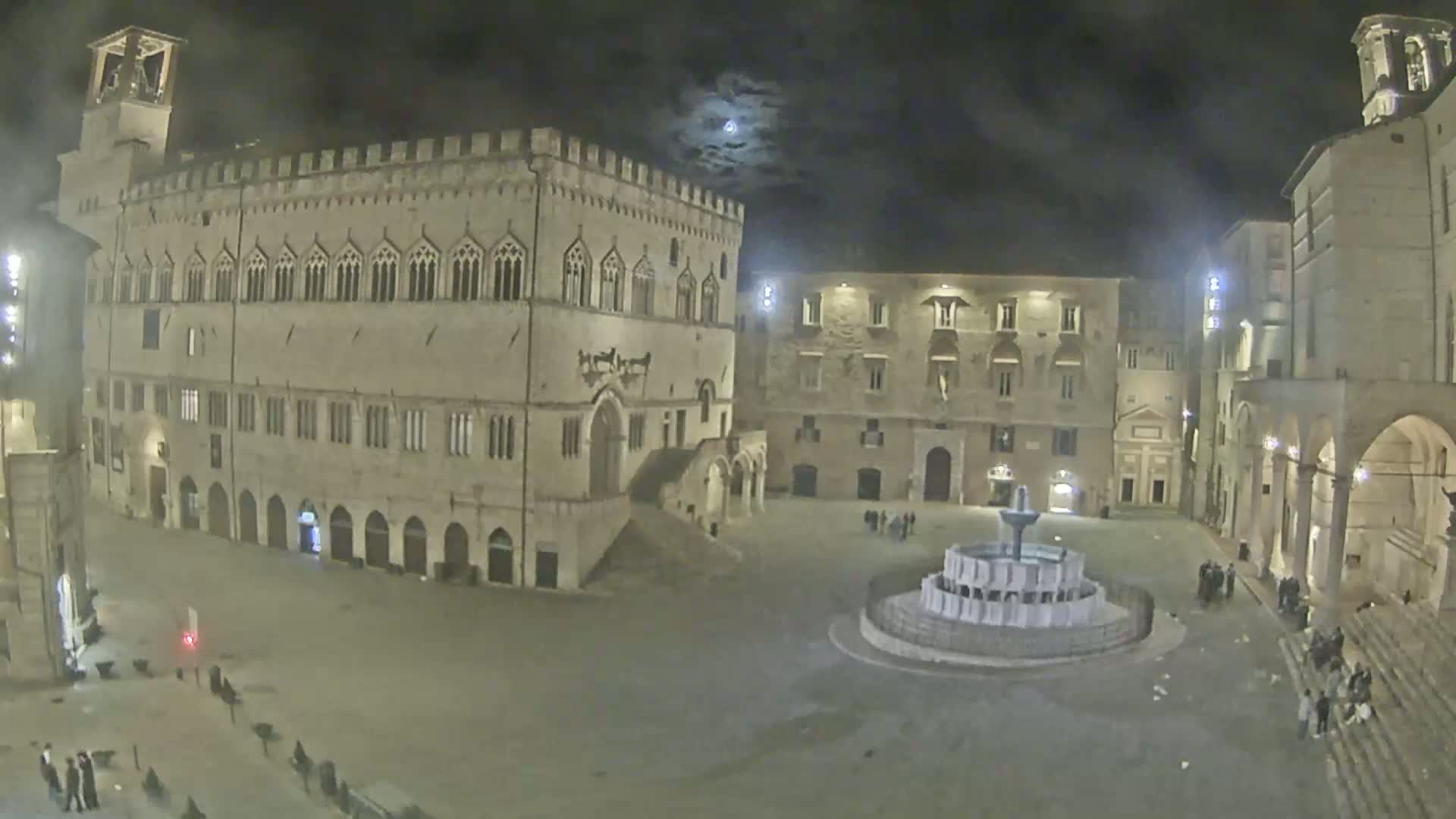 A moonlit town square at night, featuring a large stone building, several smaller buildings, and a fountain in the center.