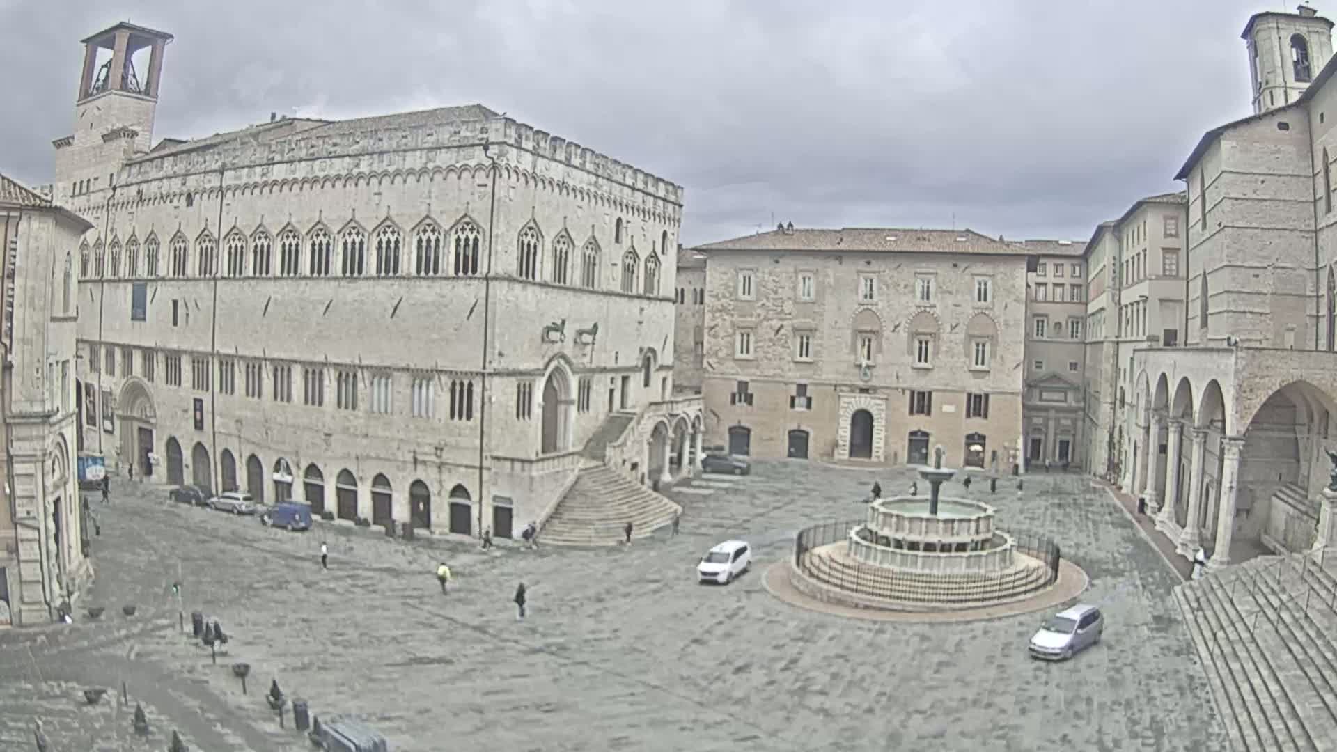 A large, stone plaza with a fountain in the center is surrounded by historic buildings under an overcast sky.
