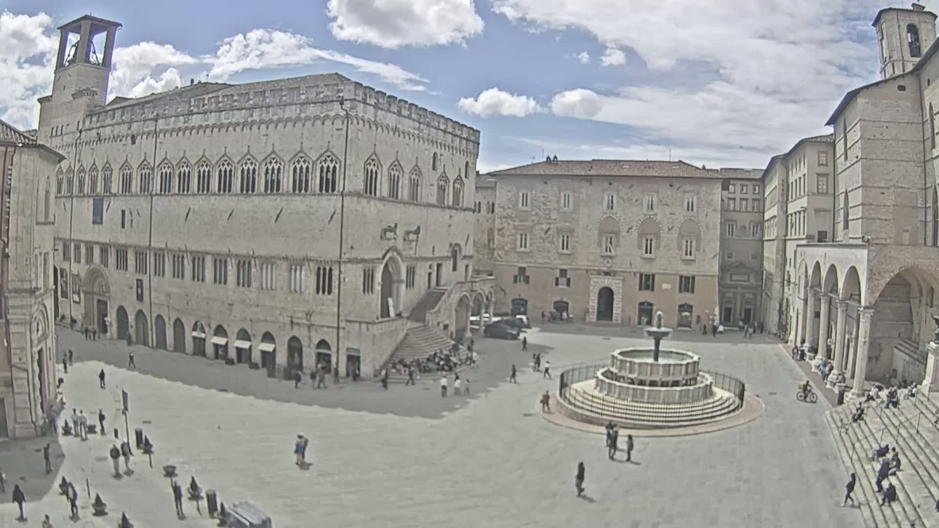 A large, light-stone town square with a fountain at its center is surrounded by historic buildings under a partly cloudy sky.