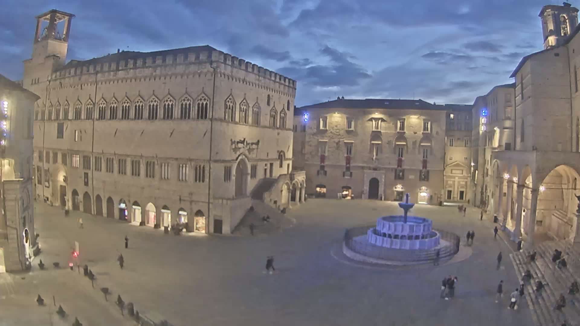 A large, sparsely populated town square at twilight, featuring several historic stone buildings and a central fountain.