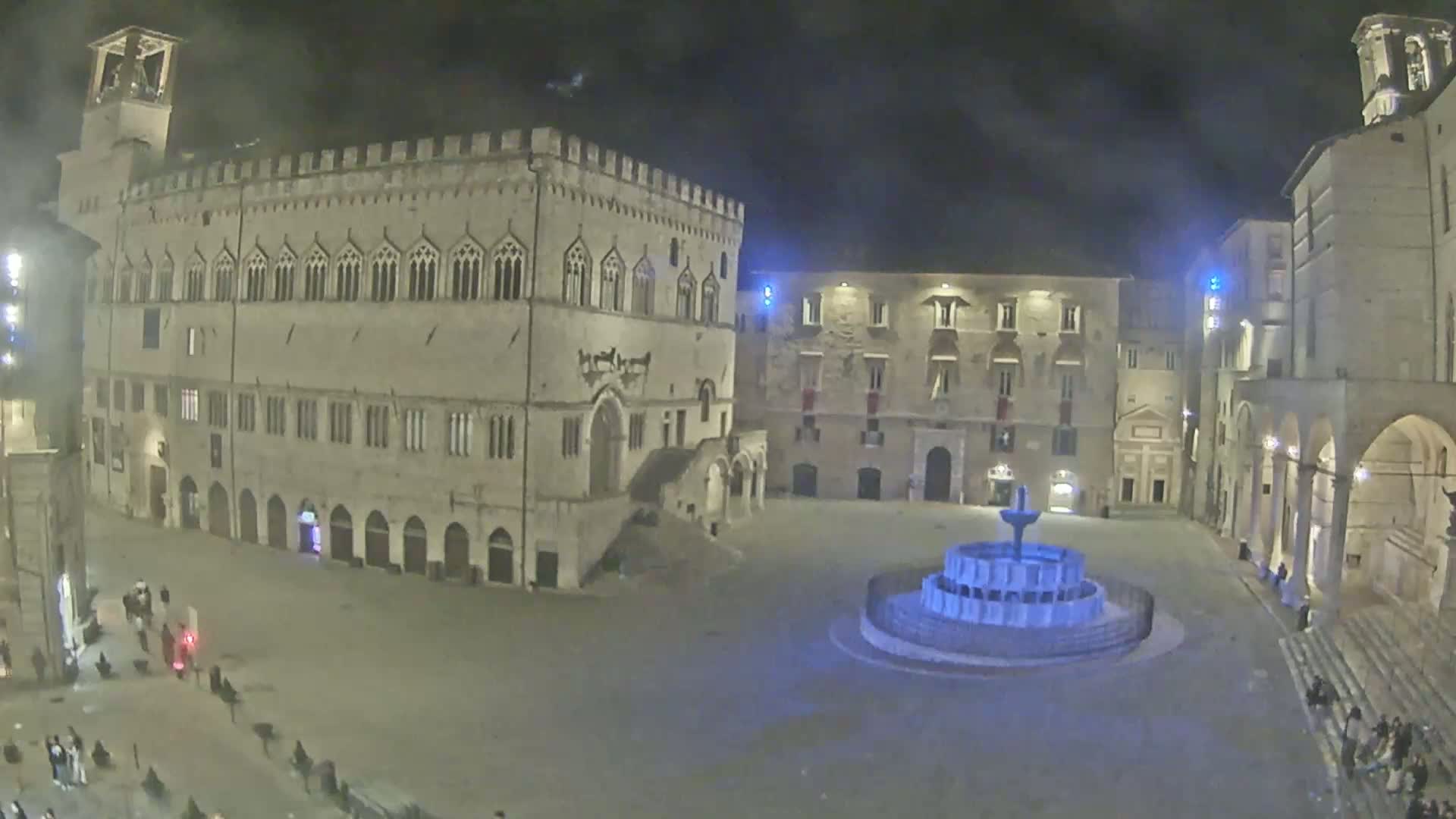 A nighttime view of a town square with a fountain, surrounded by historical stone buildings under a cloudy sky.