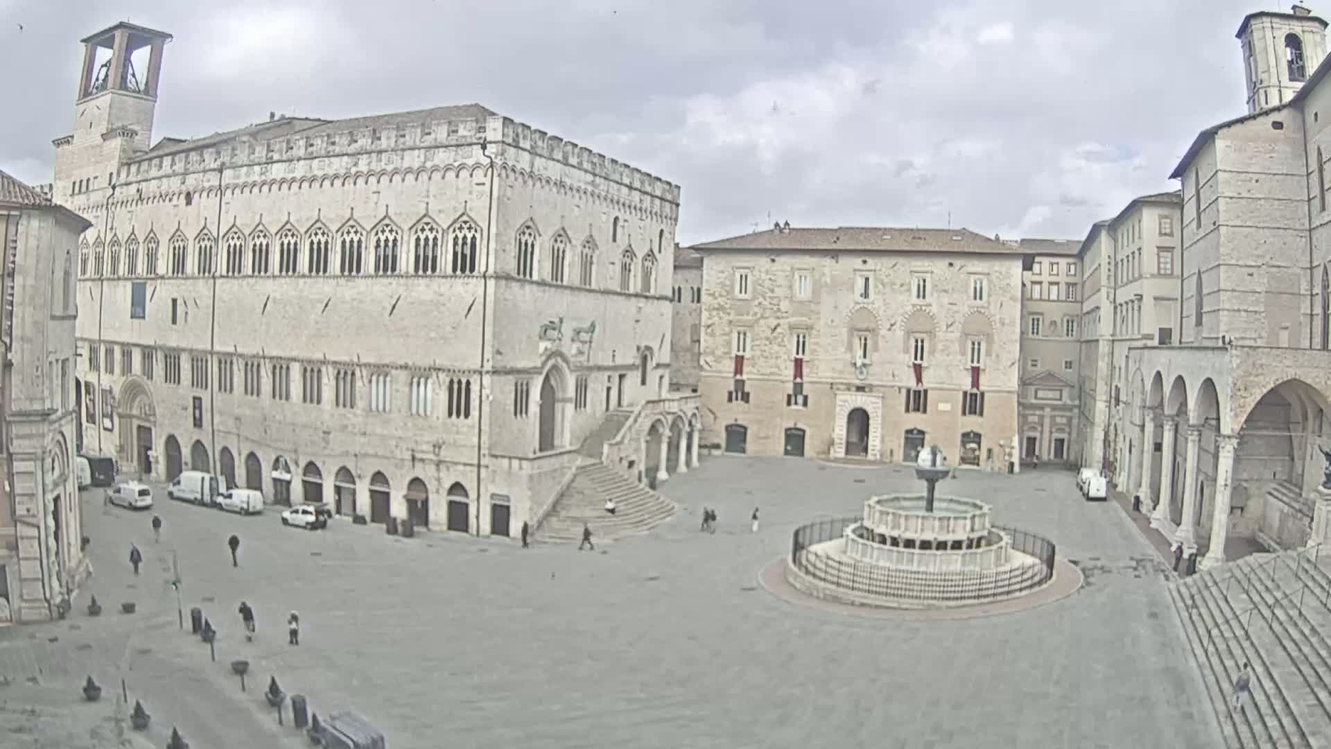 A large, mostly empty, stone plaza is centered around a fountain, with several large stone buildings surrounding it under an overcast sky.