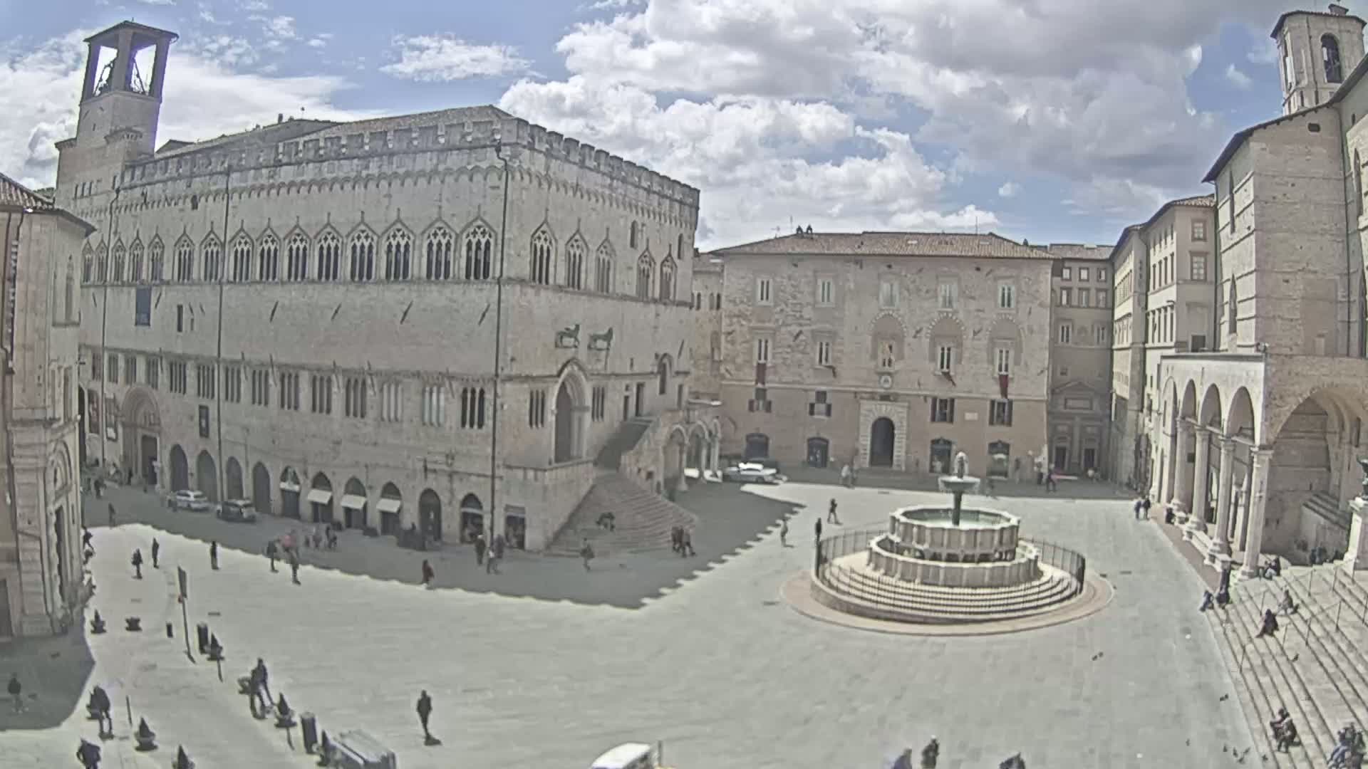 A large, light-stone town square, under a partly cloudy sky, features a central fountain and is surrounded by several large, multi-story buildings.