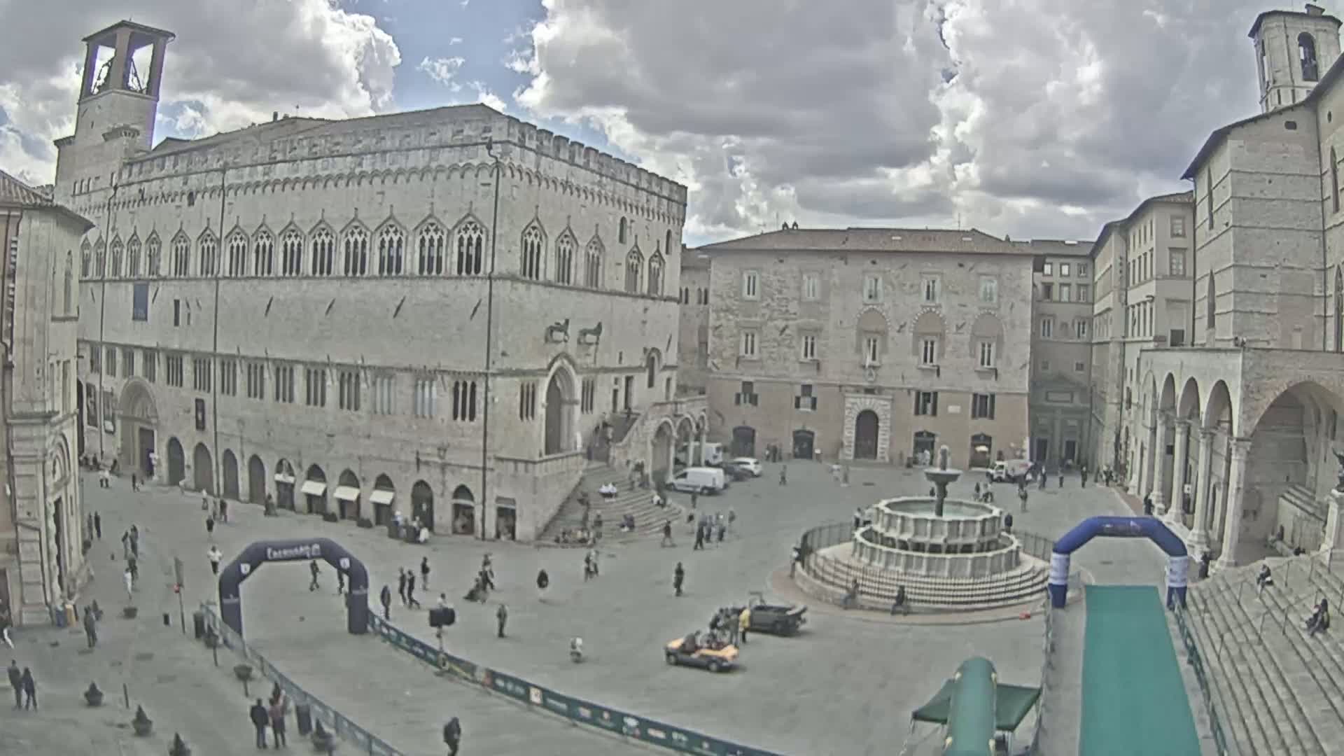 A town square with several large stone buildings, a fountain, and a few people and vehicles under a partly cloudy sky.