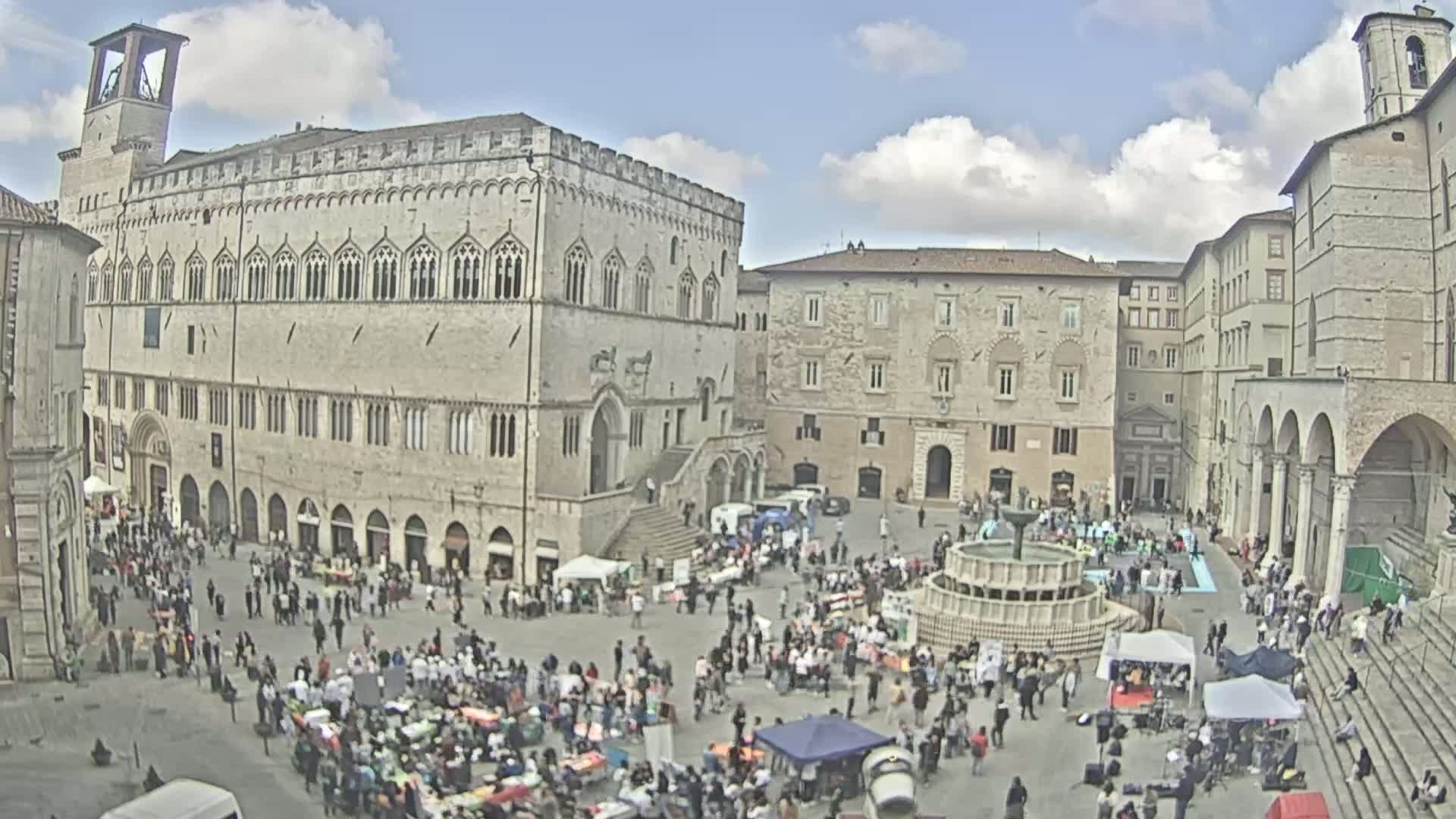 A large, crowded town square, under partly cloudy skies, is surrounded by historic stone buildings, one of which is a grand palace, and features a fountain in the center.