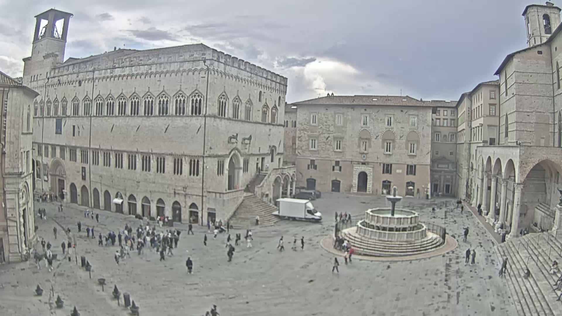A large town square with a fountain at its center is surrounded by several large stone buildings under an overcast sky.