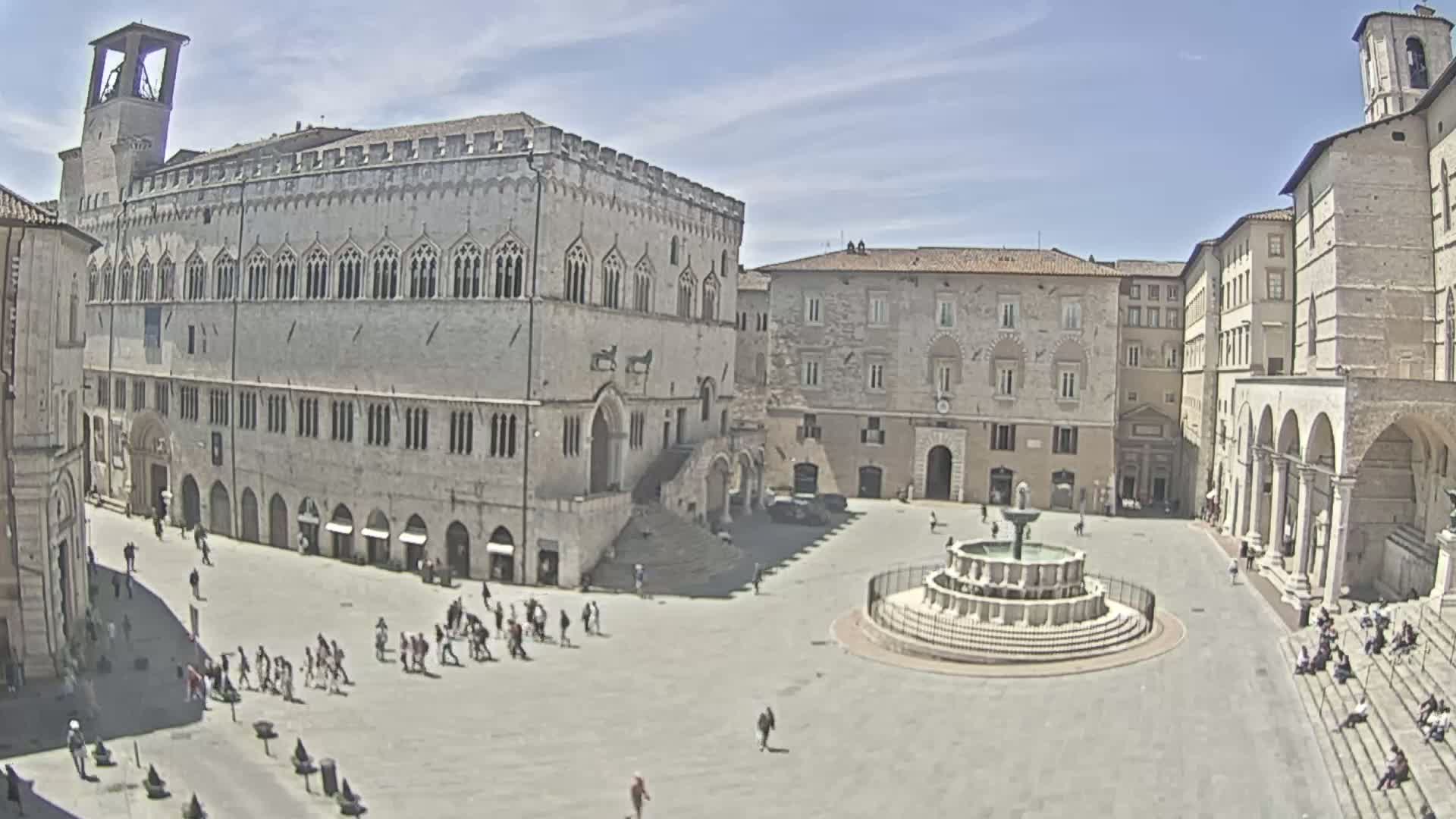 A large town square with a fountain at its center is surrounded by several stone buildings under a clear, sunny sky.