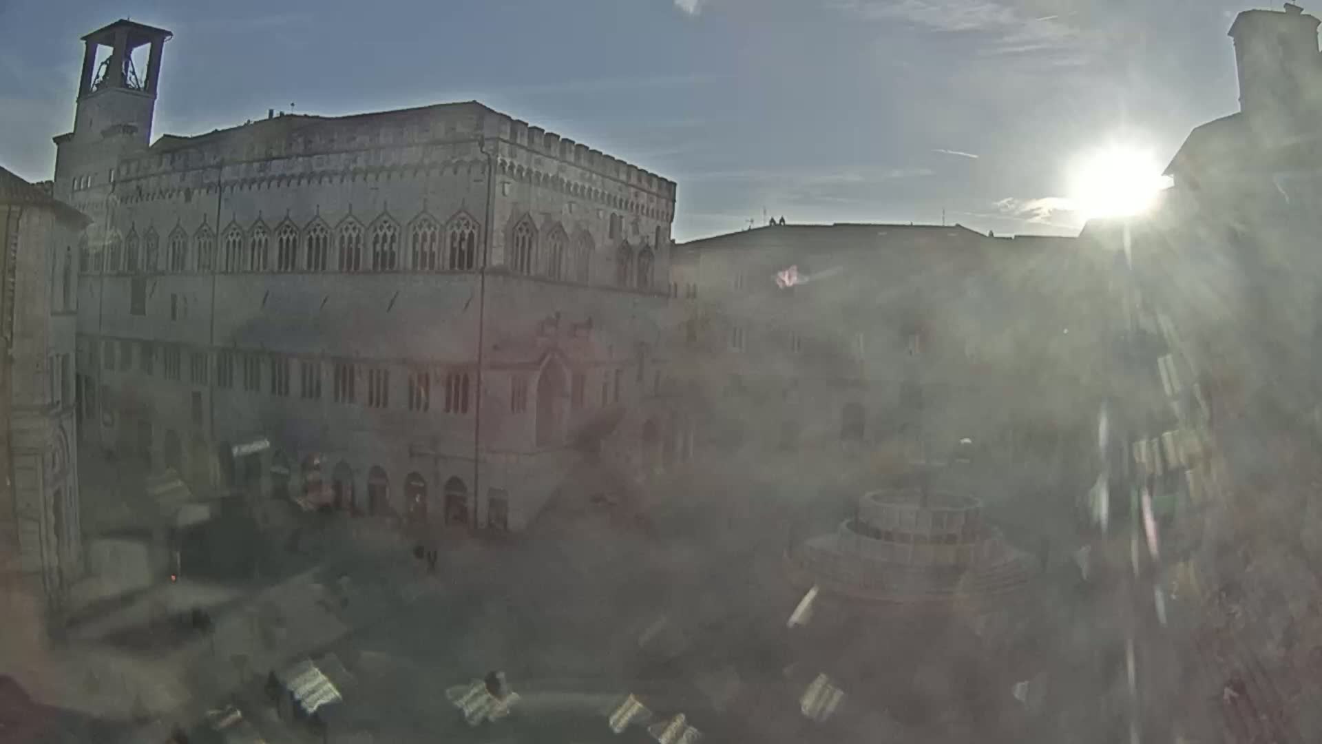 A large stone building with many windows overlooks a town square with a fountain, under a bright, hazy sun.