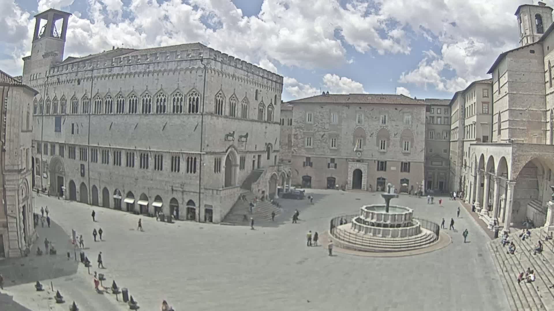 A large, mostly empty town square with a fountain at its center is surrounded by stone buildings under a partly cloudy sky.