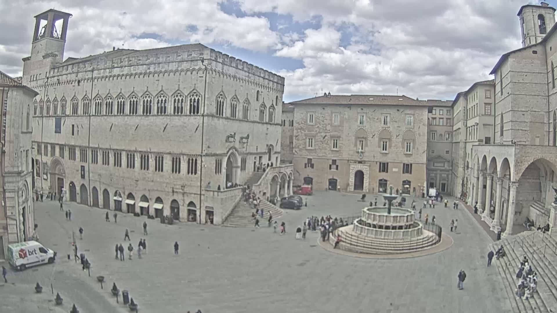 A large, mostly empty town square with a fountain at its center is surrounded by several multi-story stone buildings under a partly cloudy sky.