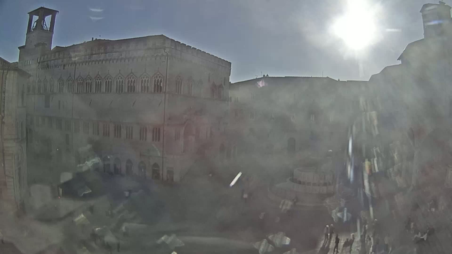 A hazy sun shines brightly over a large, stone plaza surrounded by historic buildings, one prominently featuring a bell tower.