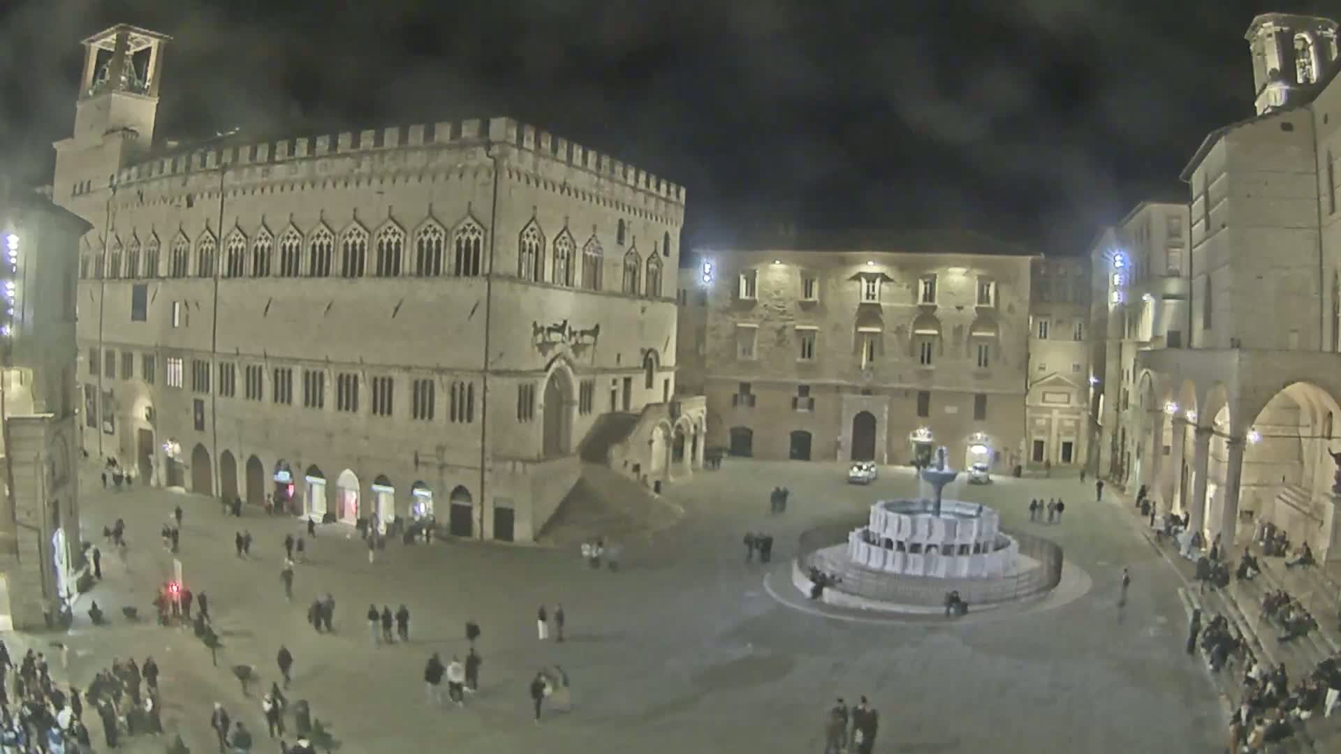A nighttime, overcast view of a large, open town square with a fountain in the center, surrounded by historic stone buildings and people.