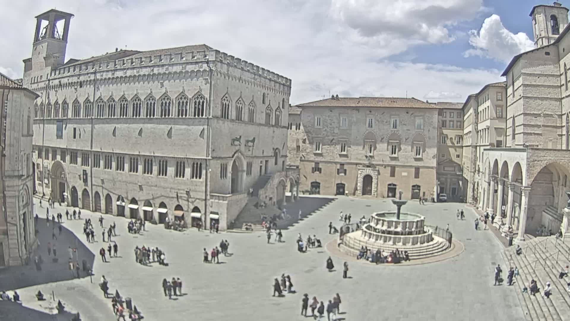 A large town square with a fountain in the center, surrounded by historic stone buildings under a partly cloudy sky.