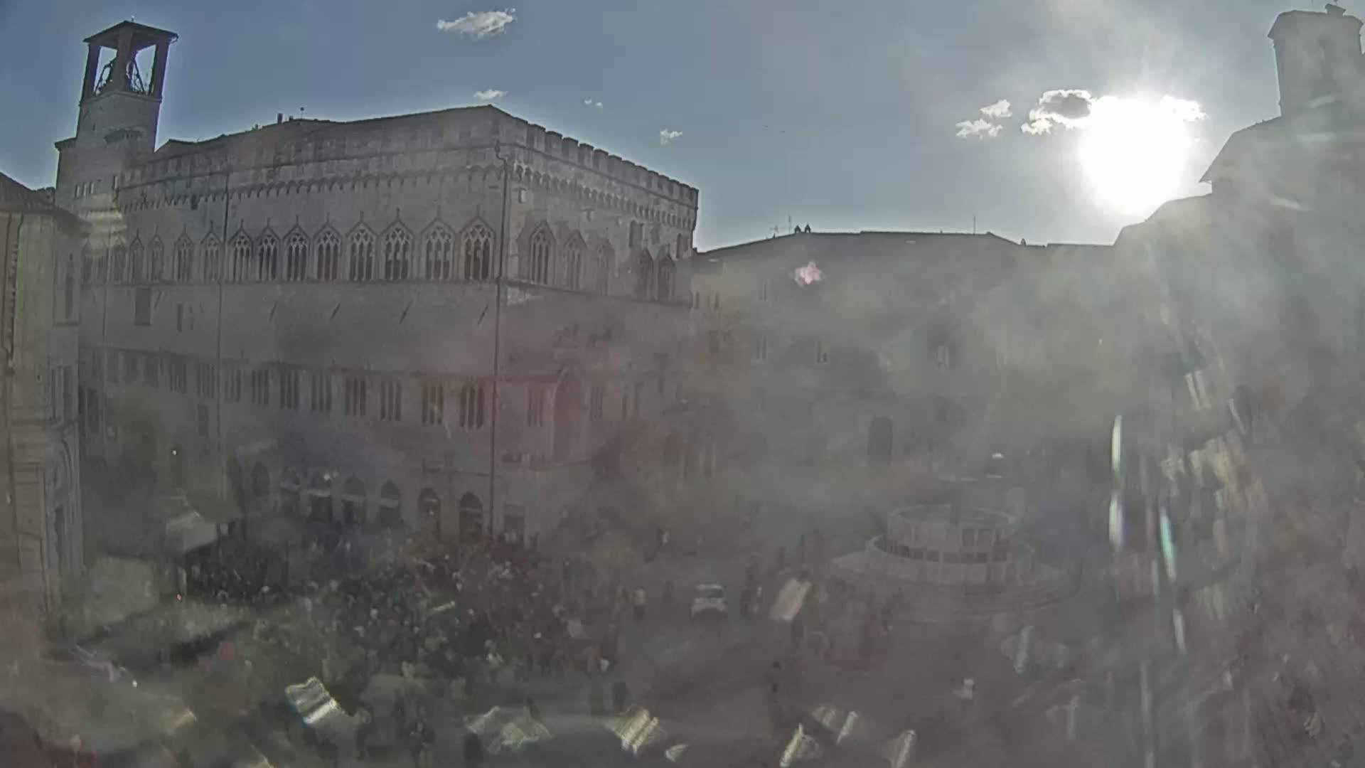 A sun-drenched, hazy view of a large, crowded town square dominated by a tall, stone building with many windows.