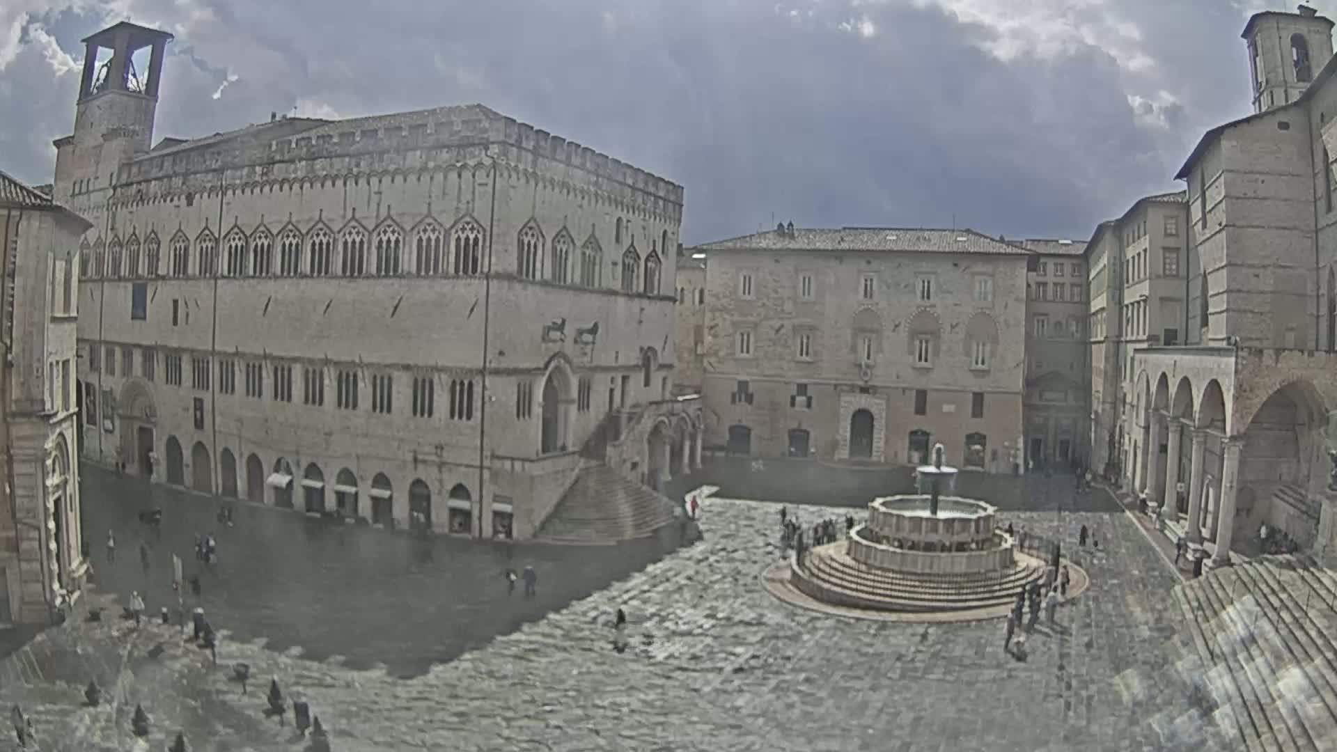 A large, stone plaza with a fountain is surrounded by several medieval-style buildings under a cloudy sky.