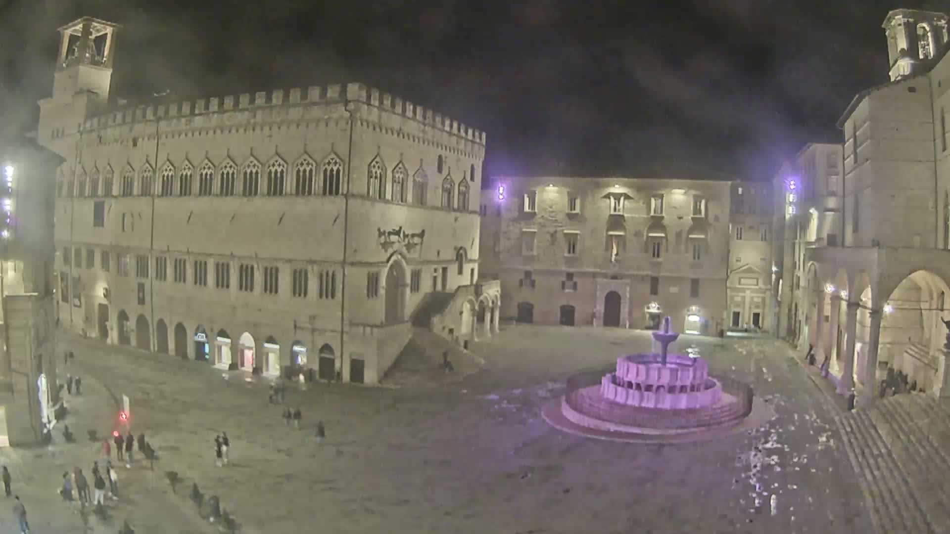 A nighttime view of a town square with a fountain, surrounded by historic stone buildings under a cloudy sky.