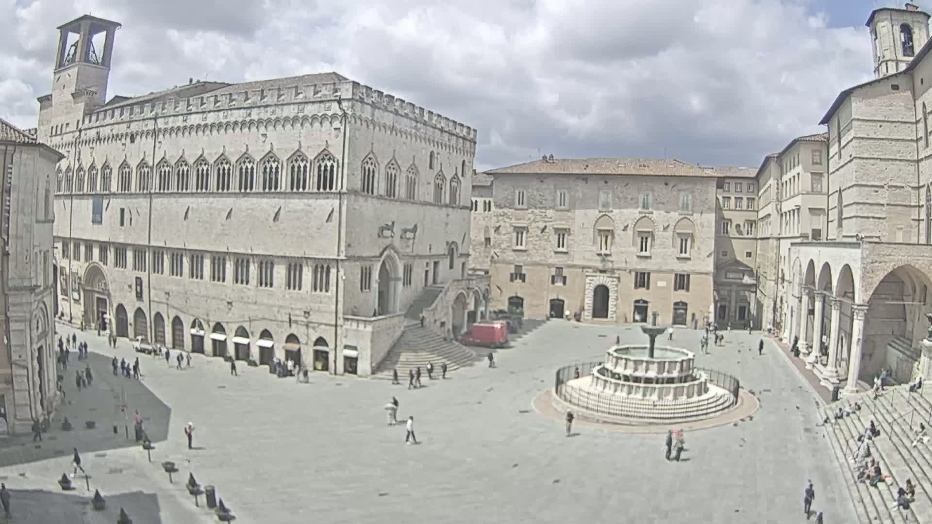 A large, mostly empty town square with a fountain in the center is surrounded by aged stone buildings under a partly cloudy sky.
