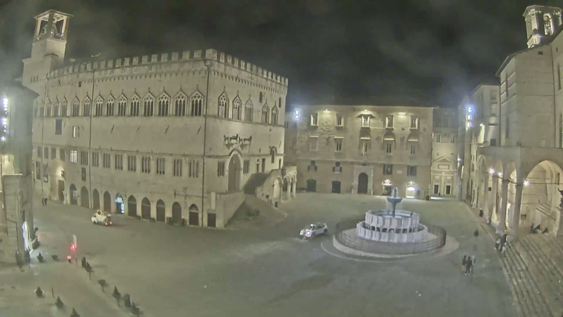 A nighttime view of a large, mostly empty town square with a fountain at its center, surrounded by historic stone buildings.