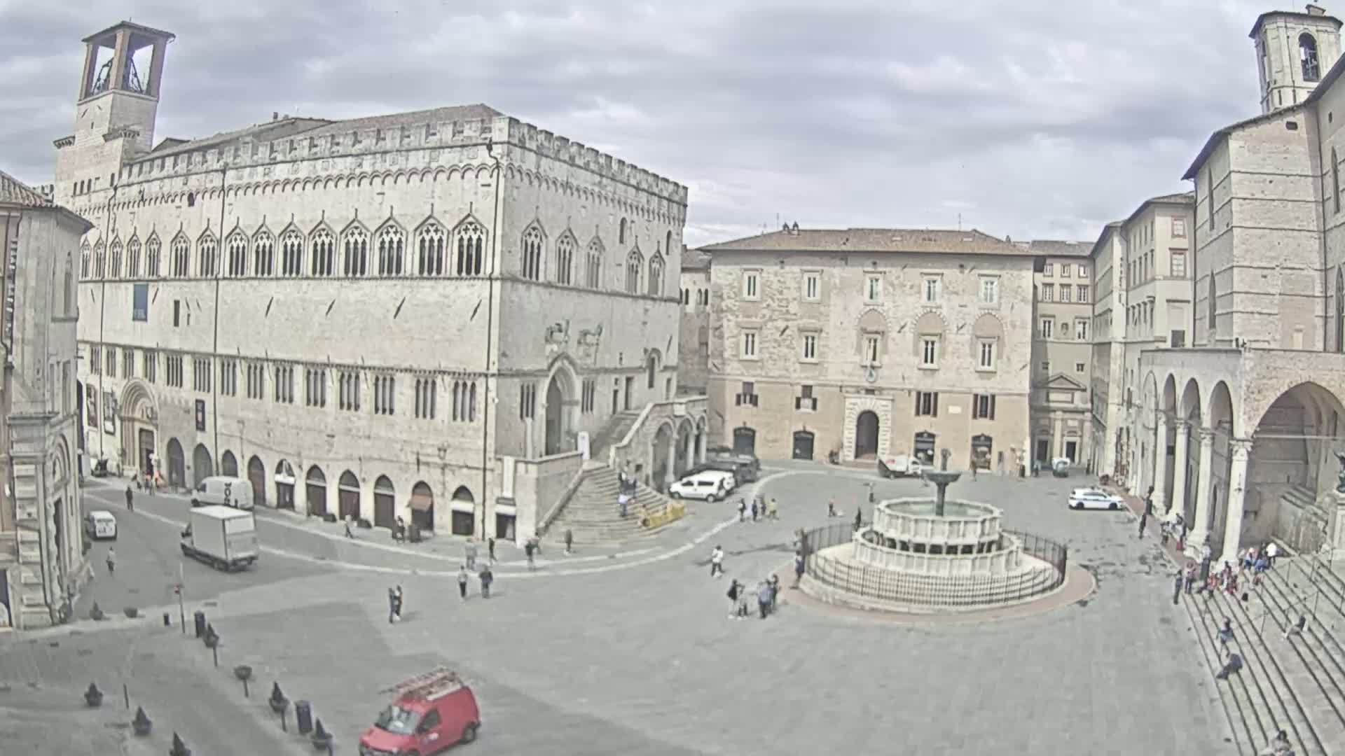A large town square with several stone buildings surrounding a central fountain, under a partly cloudy sky.