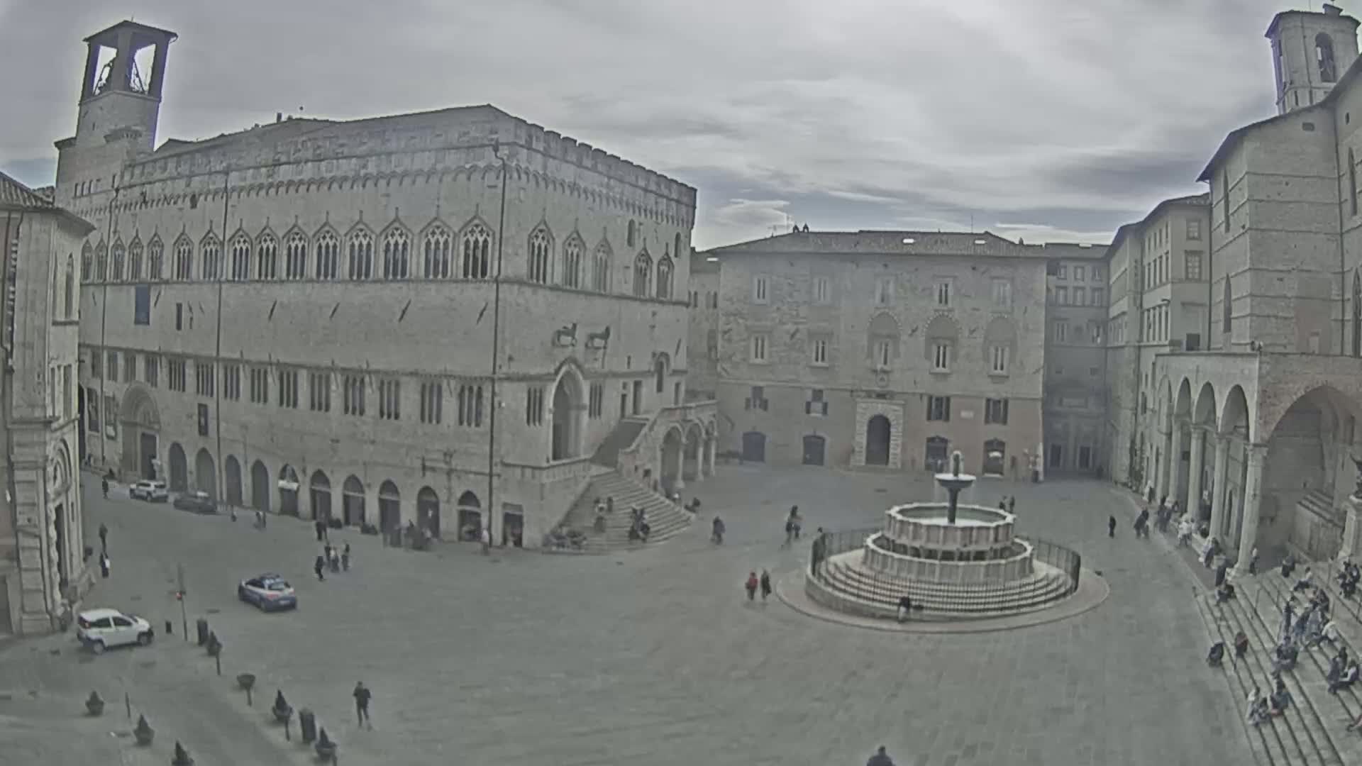 A large town square with several stone buildings surrounding a central fountain, under an overcast sky.