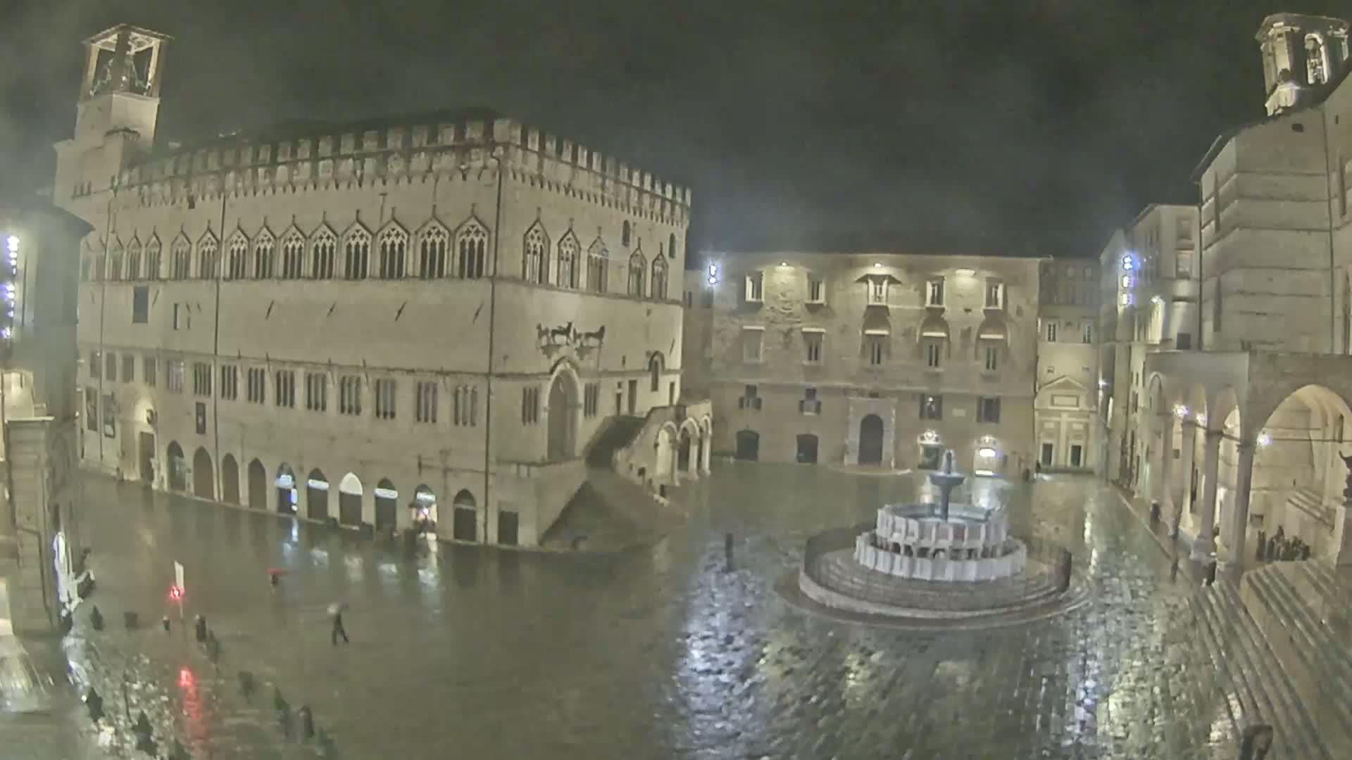 A large, stone plaza at night, illuminated by lights, is flooded with rainwater, showing a fountain in the center and several buildings surrounding it.