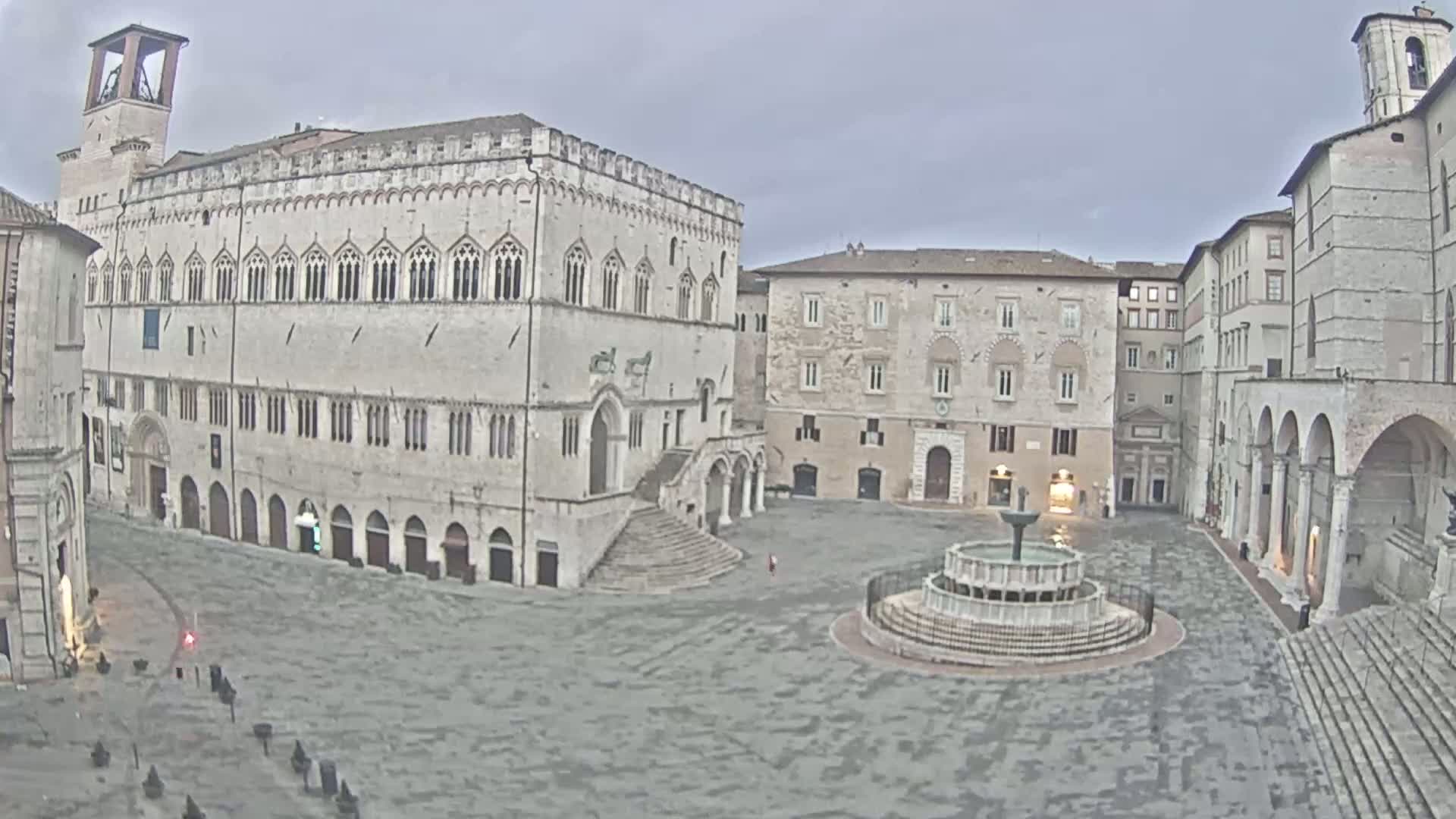 A large, mostly empty, stone plaza with a fountain at its center is surrounded by aged, multi-story buildings under an overcast sky.