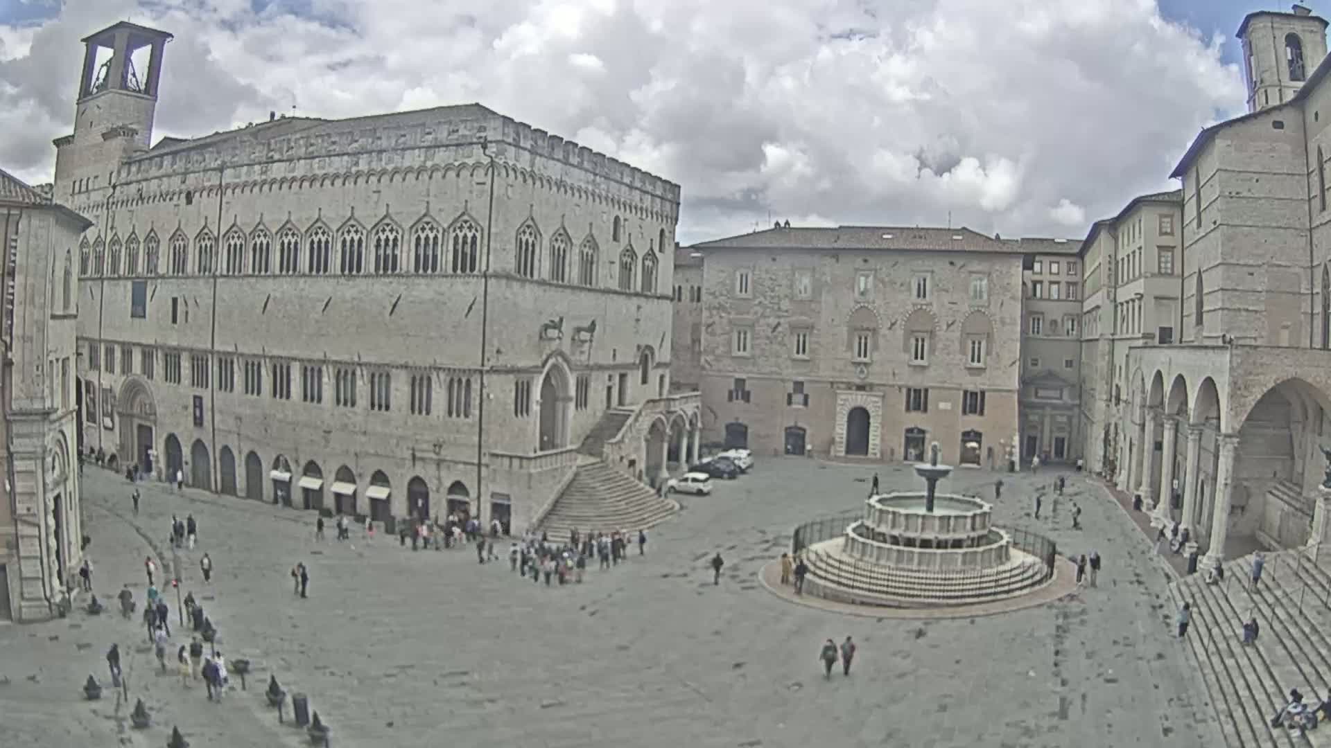A large, stone plaza with a fountain at its center, surrounded by historic buildings under a partly cloudy sky.