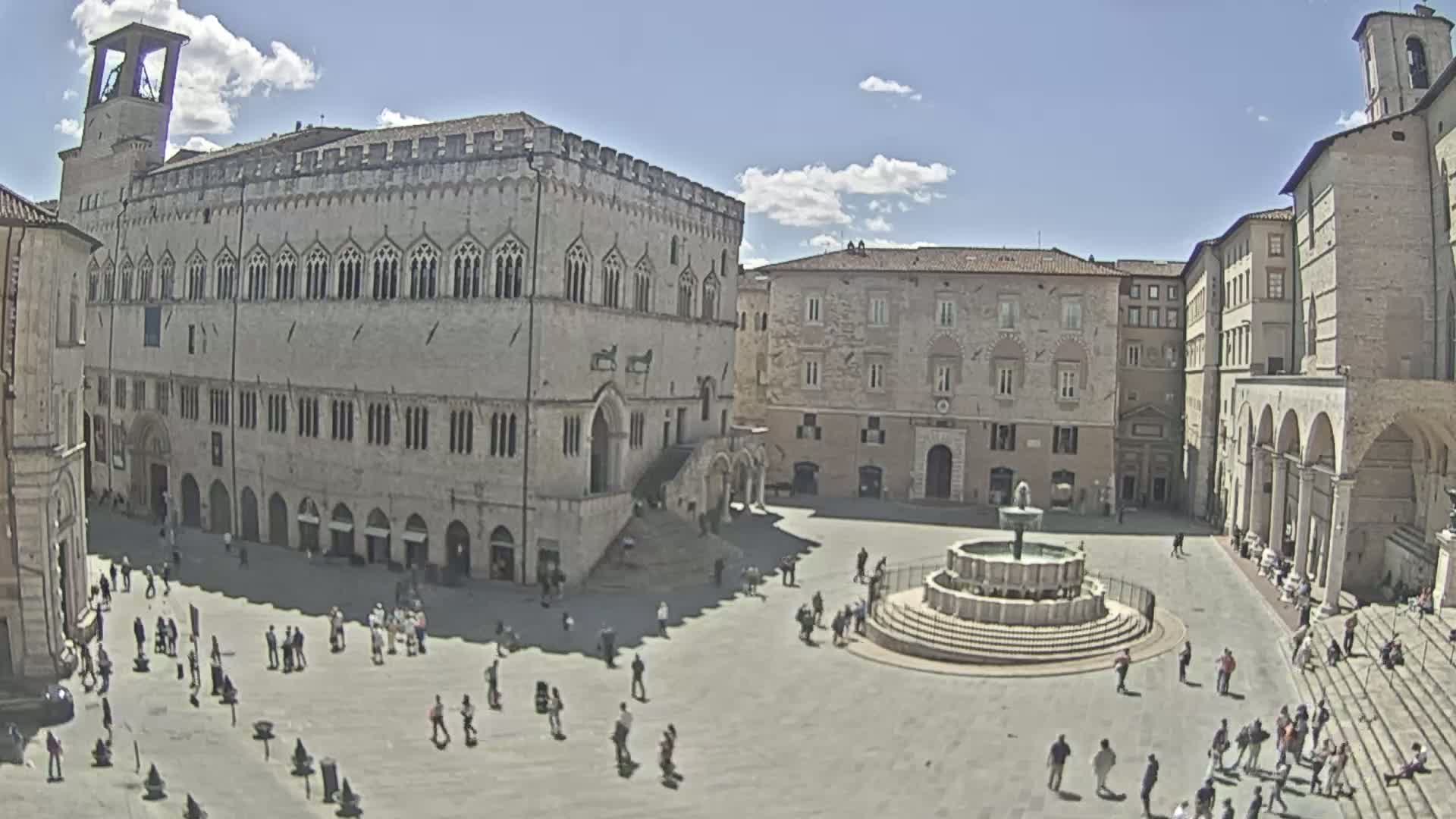A sunny day in a large, stone plaza in Italy, featuring a large building with arched windows, a fountain in the center, and many people scattered throughout.