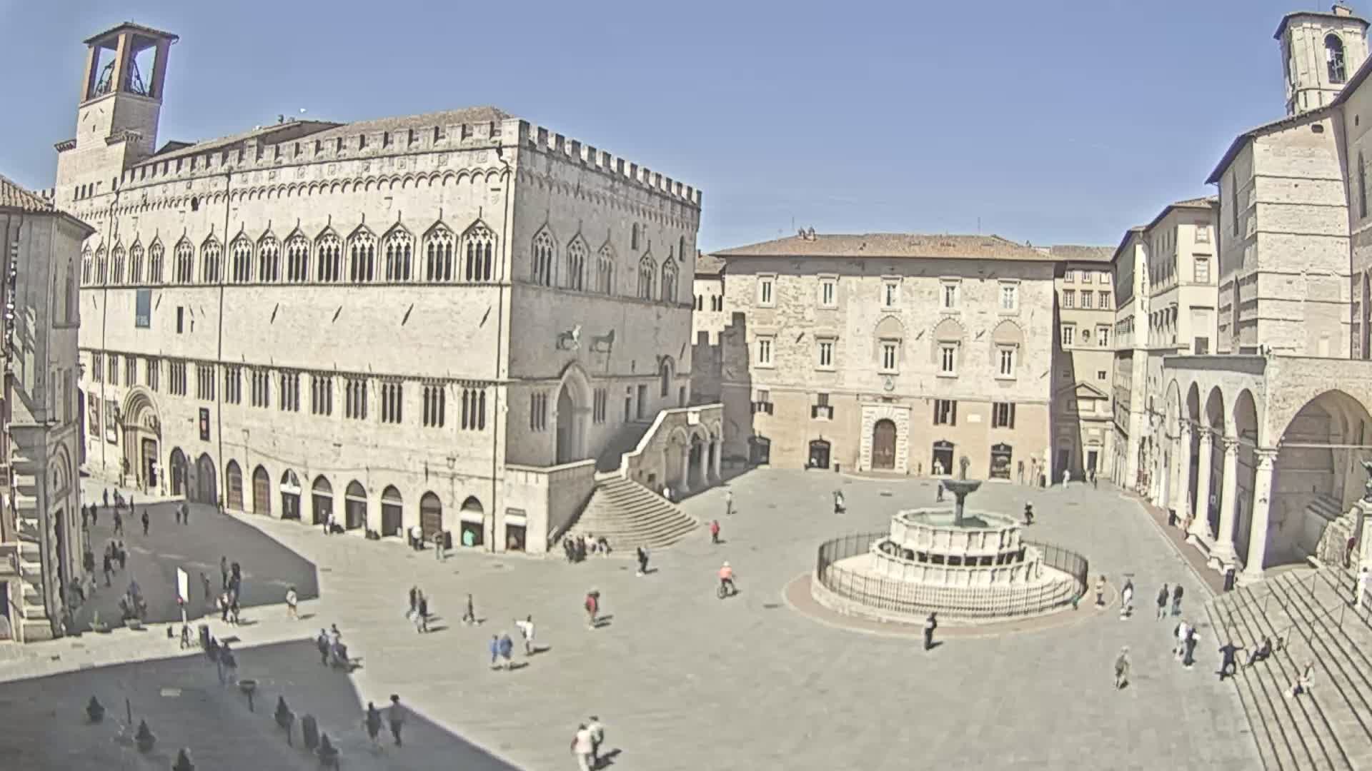 A large town square with stone buildings, a fountain in the center, and a few people scattered throughout, under a sunny sky.
