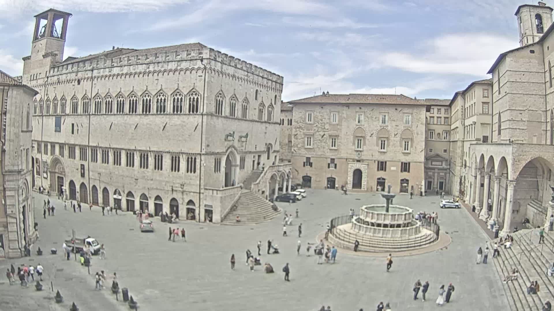 A large town square with several stone buildings, a fountain, and a scattering of people under partly cloudy skies.