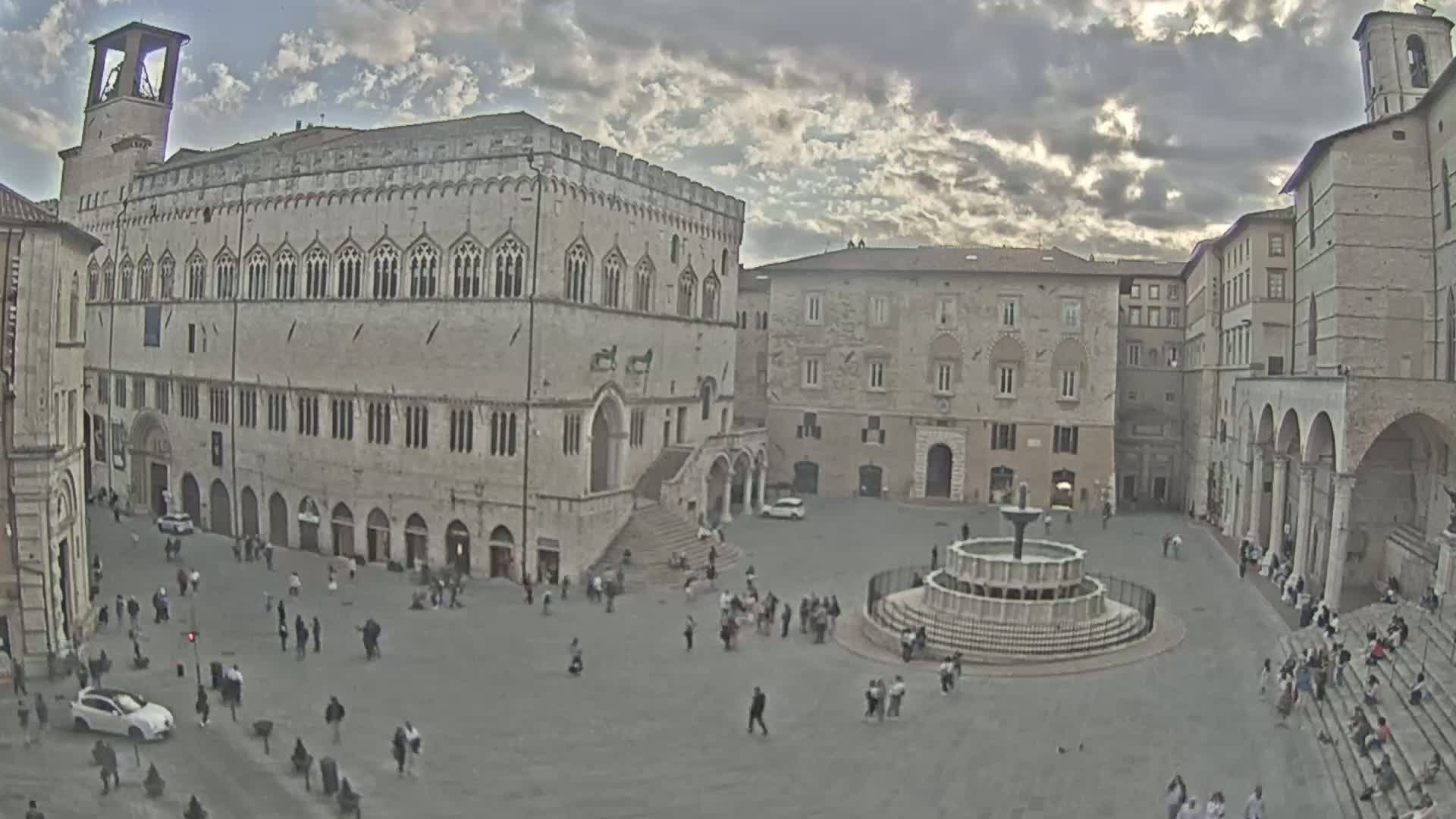 A large town square with a fountain is surrounded by stone buildings under a partly cloudy sky.