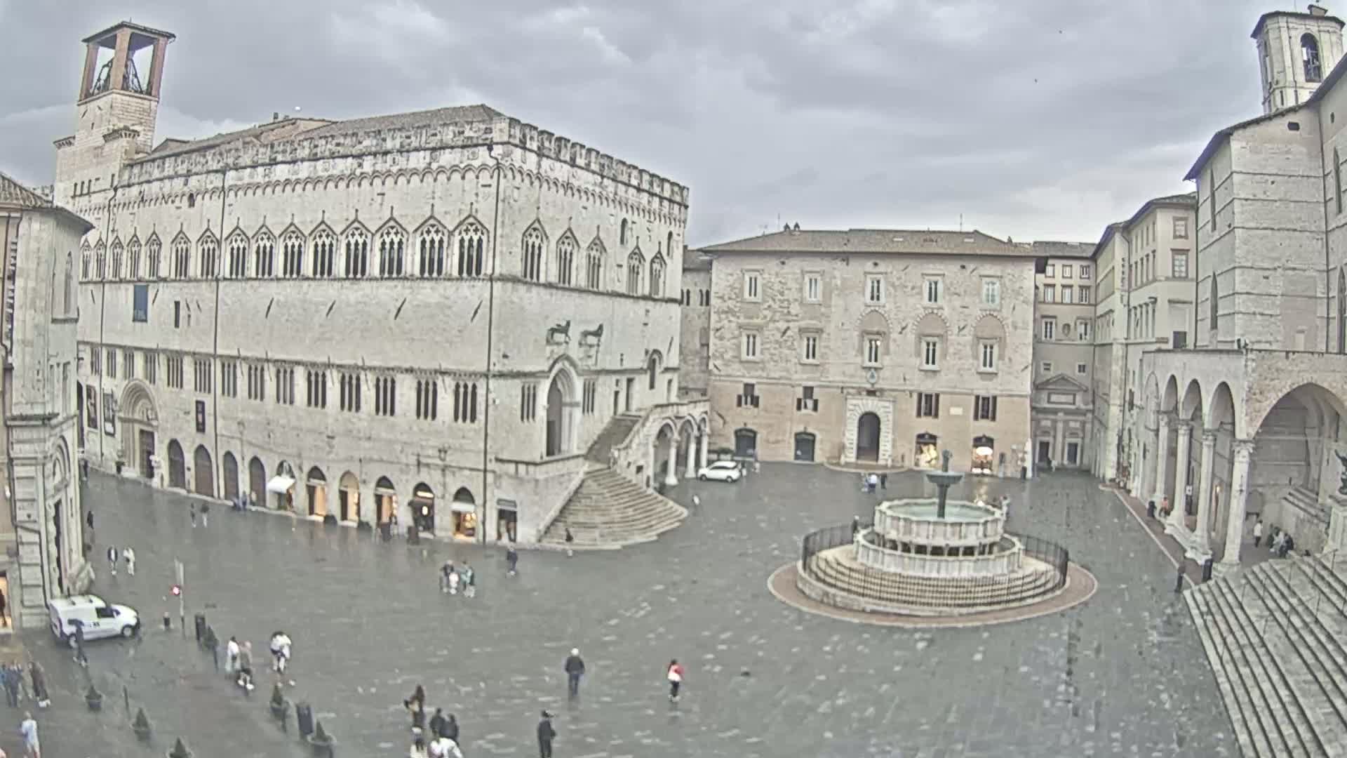 A large town square with several stone buildings, a fountain, and a few pedestrians, under an overcast sky.