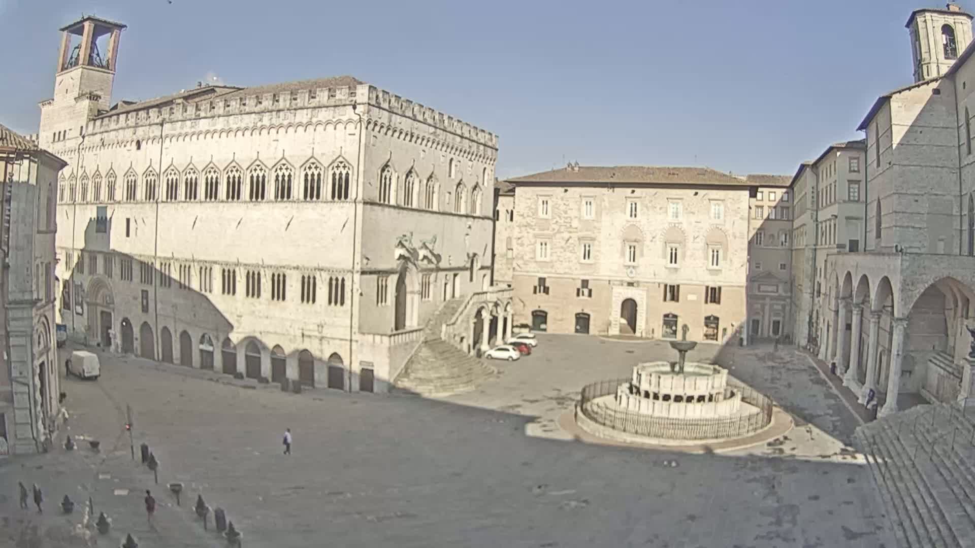 A large, mostly empty town square is centered around a fountain, flanked by several large, light-colored stone buildings under a bright, sunny sky.
