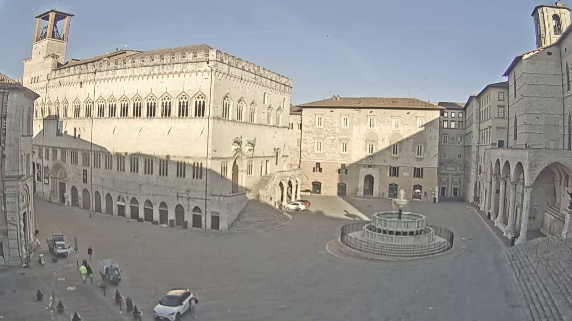 A large, mostly empty town square is centered around a fountain, surrounded by several light-colored, historic buildings under sunny skies.