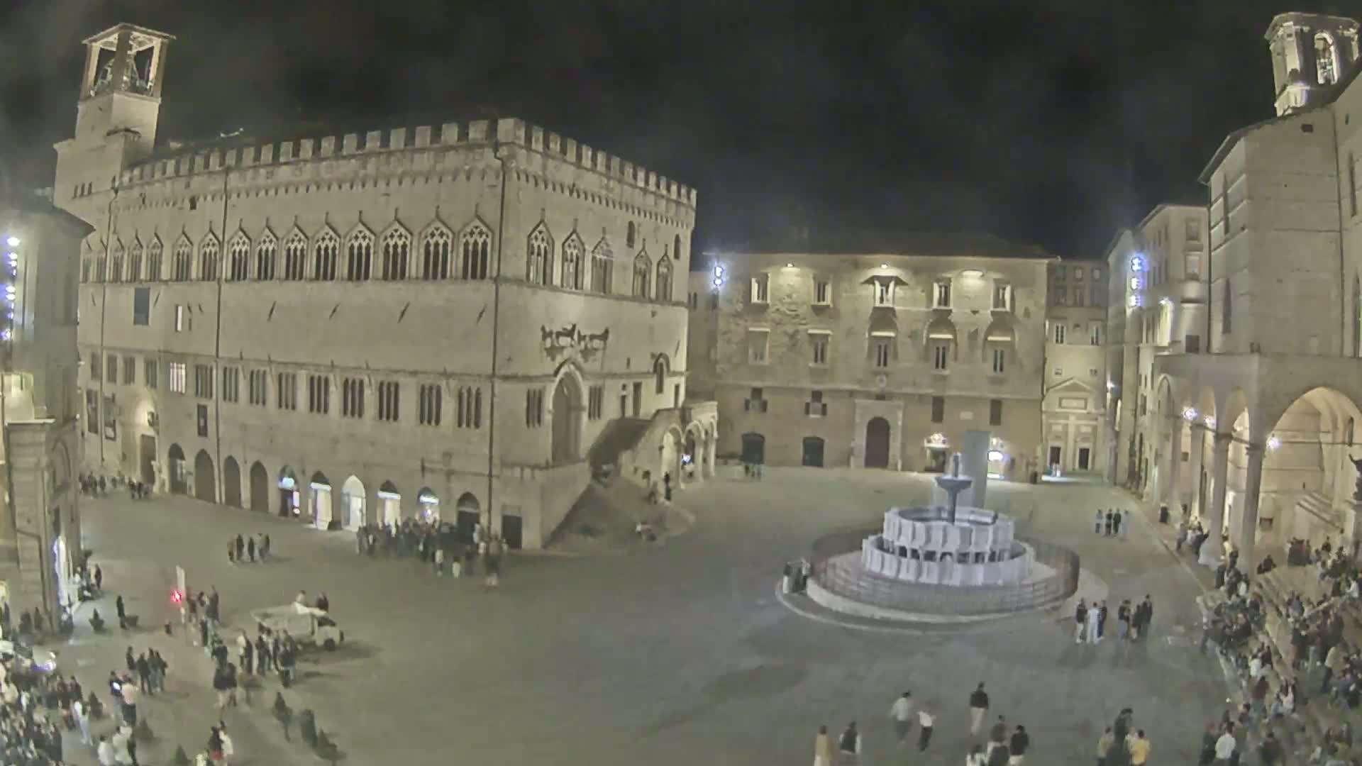 A nighttime view of a town square with a fountain, surrounded by large stone buildings and a small crowd of people.