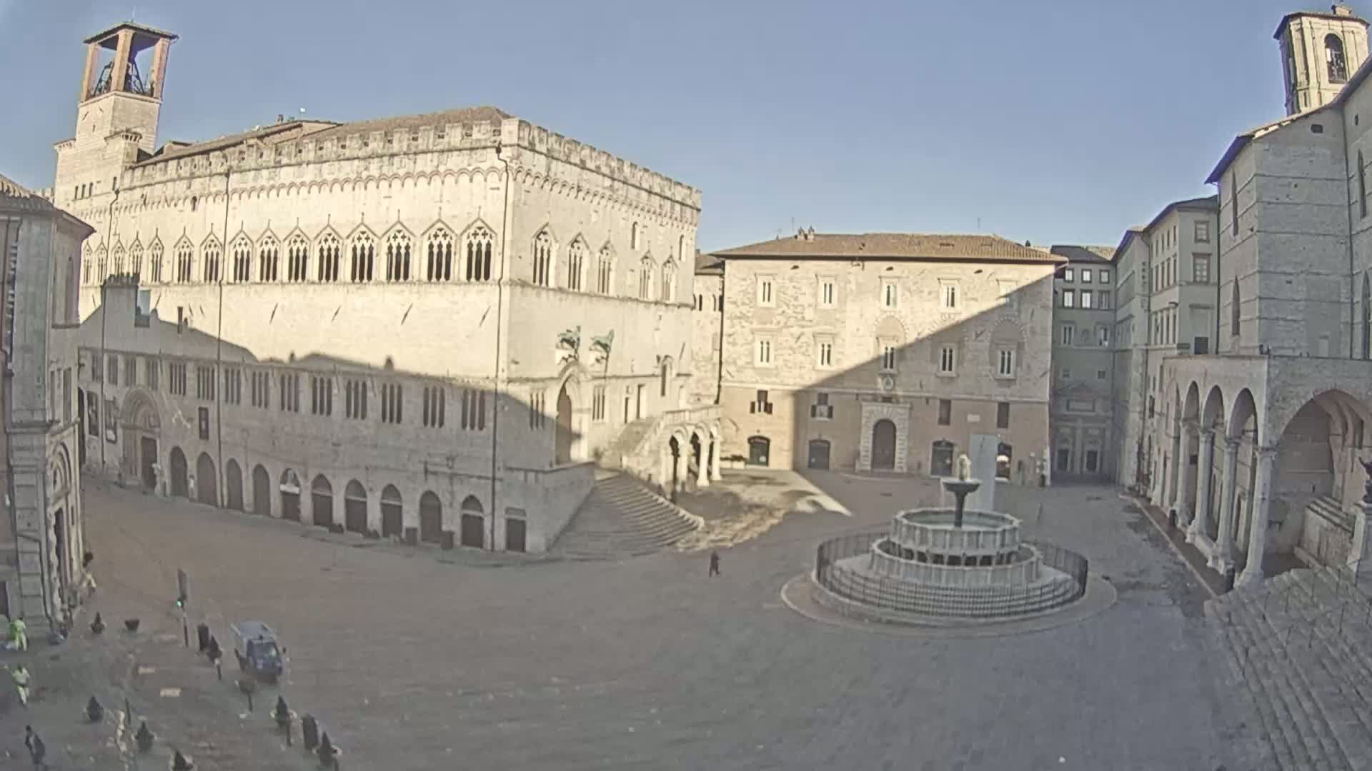 A large, mostly empty town square with several stone buildings, a fountain, and a clear sunny day.