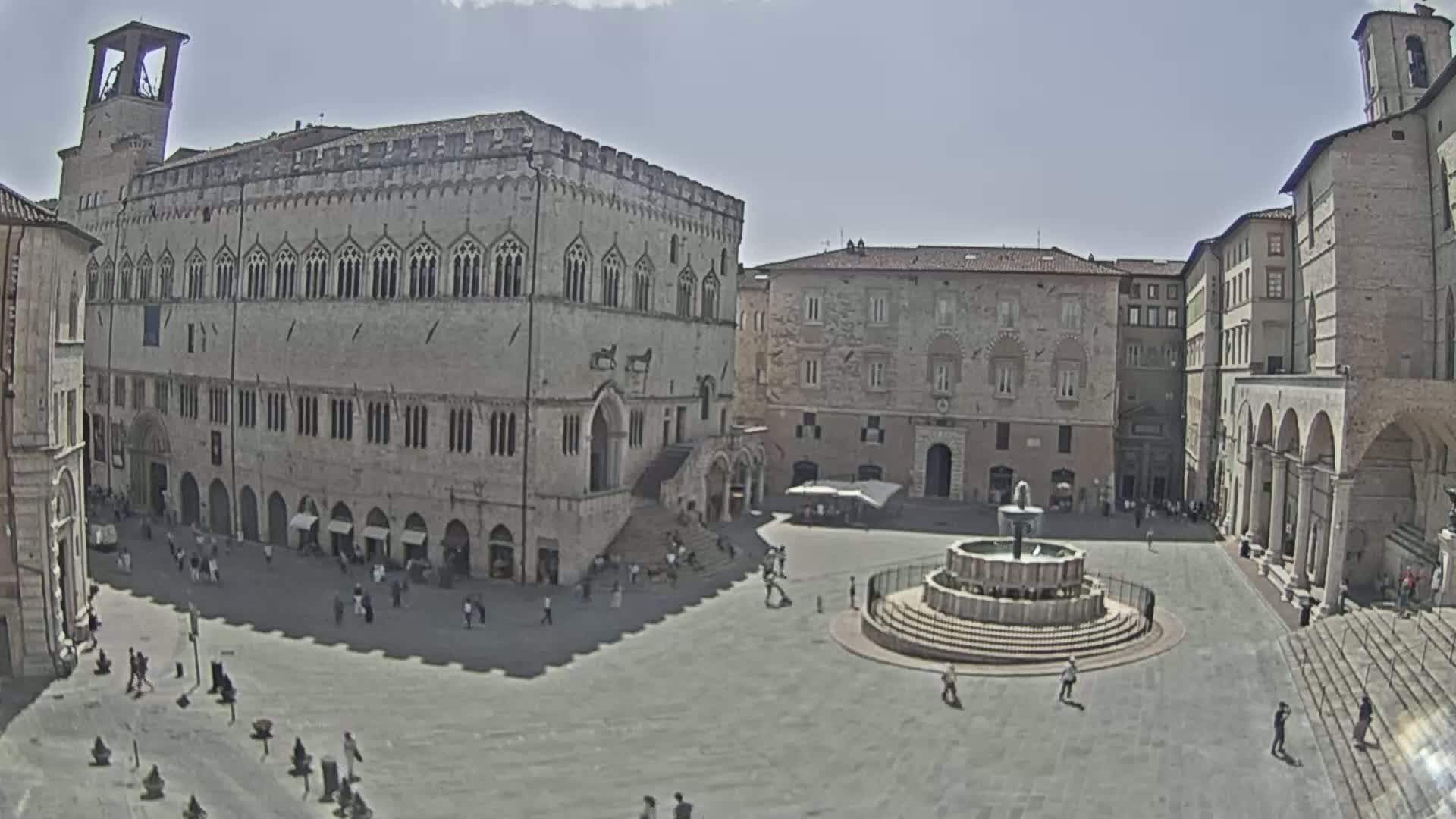 A sunny town square is shown, featuring a large stone building with arched windows and a central fountain, surrounded by other historical-looking buildings.