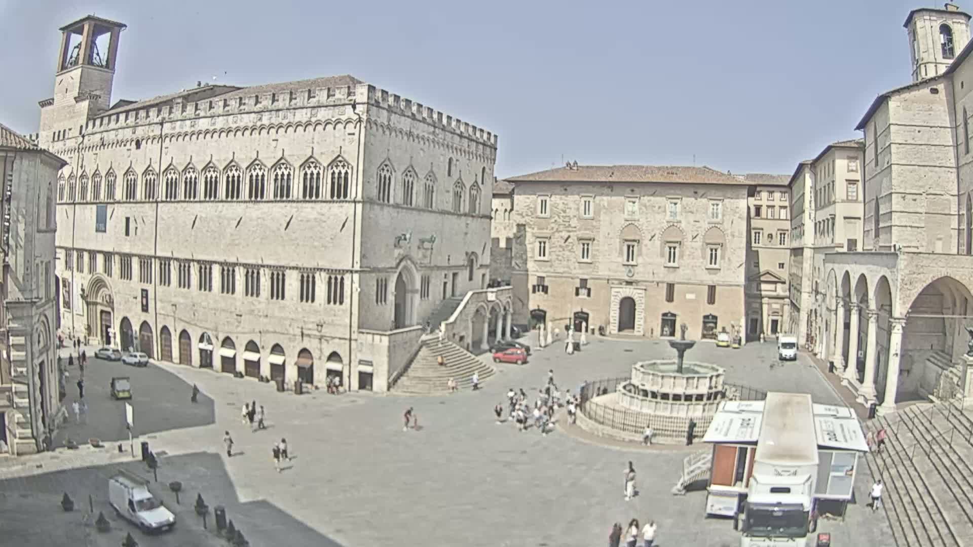 A large, sunlit town square features several stone buildings, a fountain, and a scattering of people and vehicles.