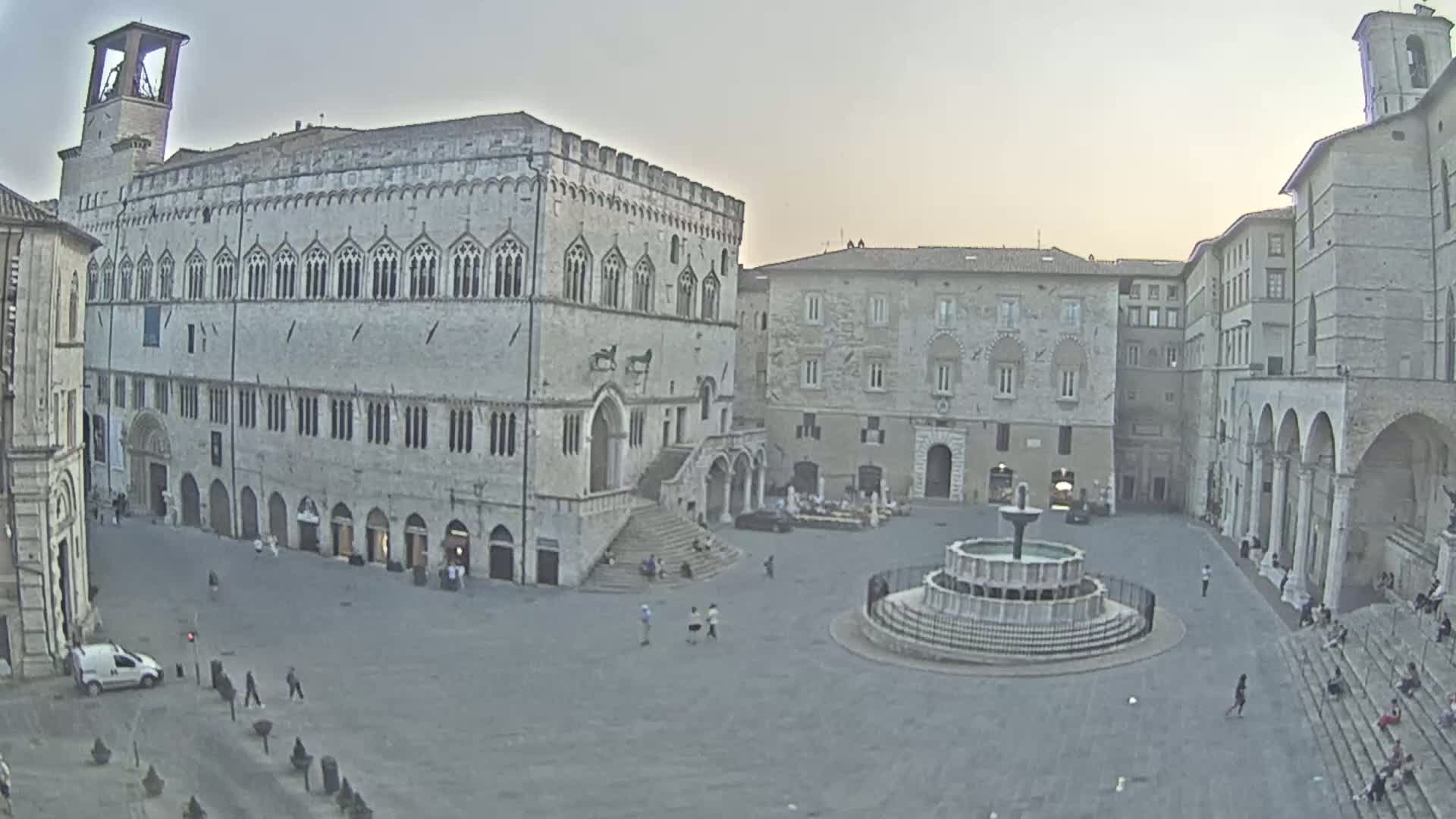 A large, stone plaza with a fountain at its center is surrounded by historical buildings under a pale, overcast sky.