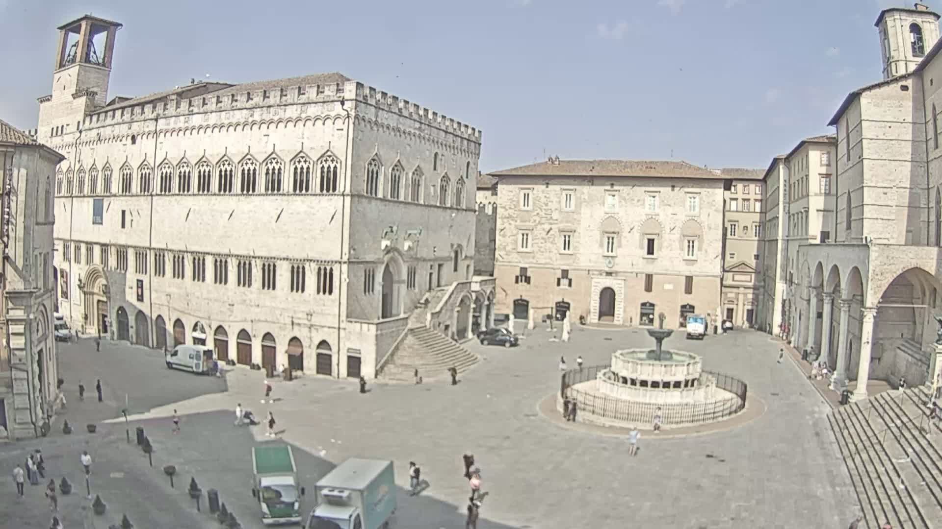 A large, sunny town square features a central fountain, surrounded by several historic stone buildings, some with arched entryways and multiple stories.