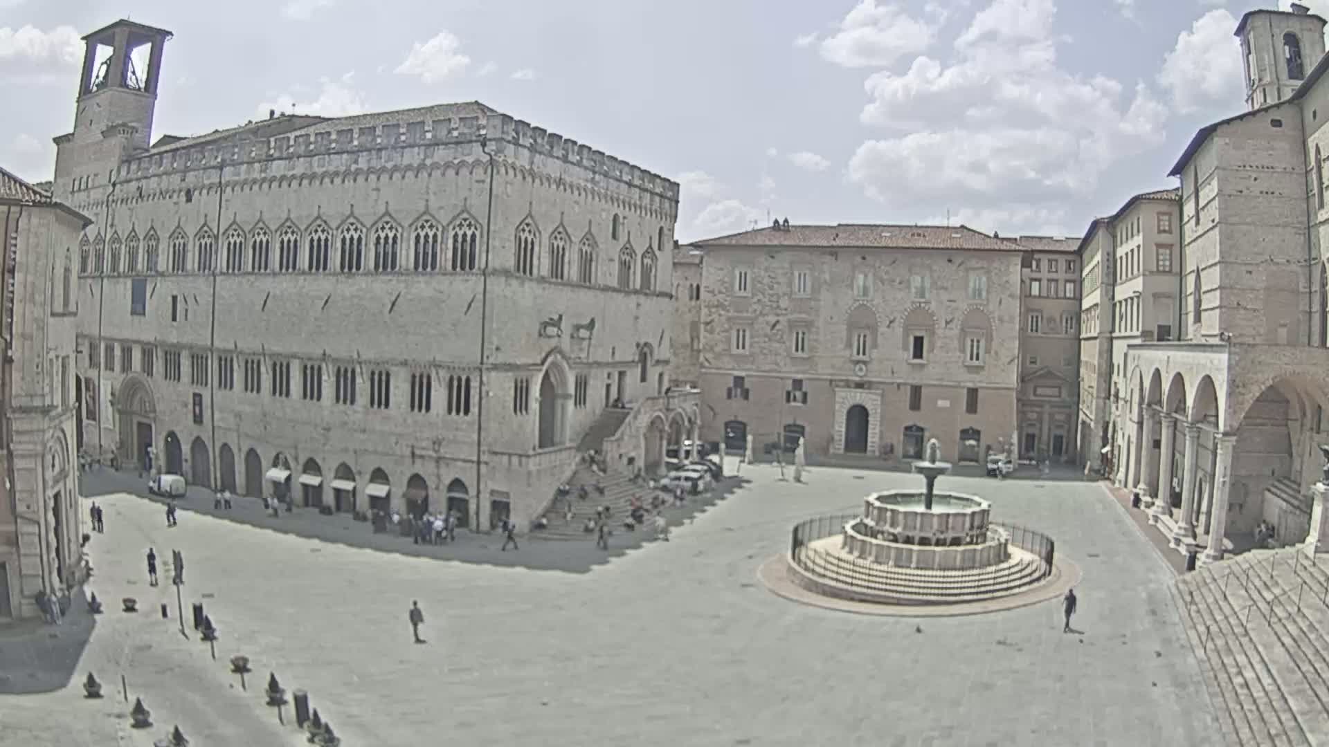 A large, stone plaza with a fountain in the center is surrounded by historic buildings under a partly cloudy sky.
