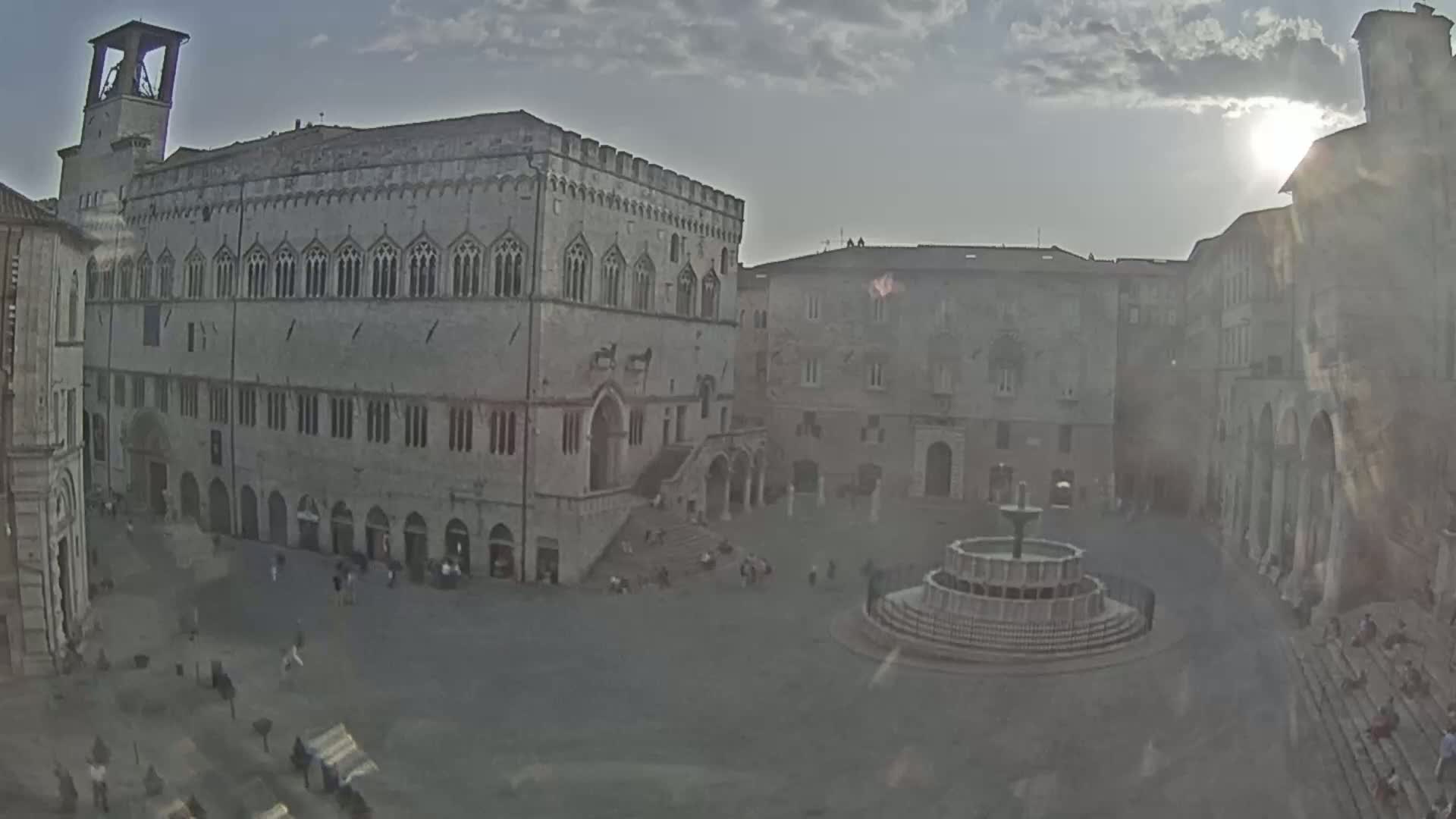 A large, stone public square with a fountain at its center is surrounded by historic buildings under a partly sunny sky.