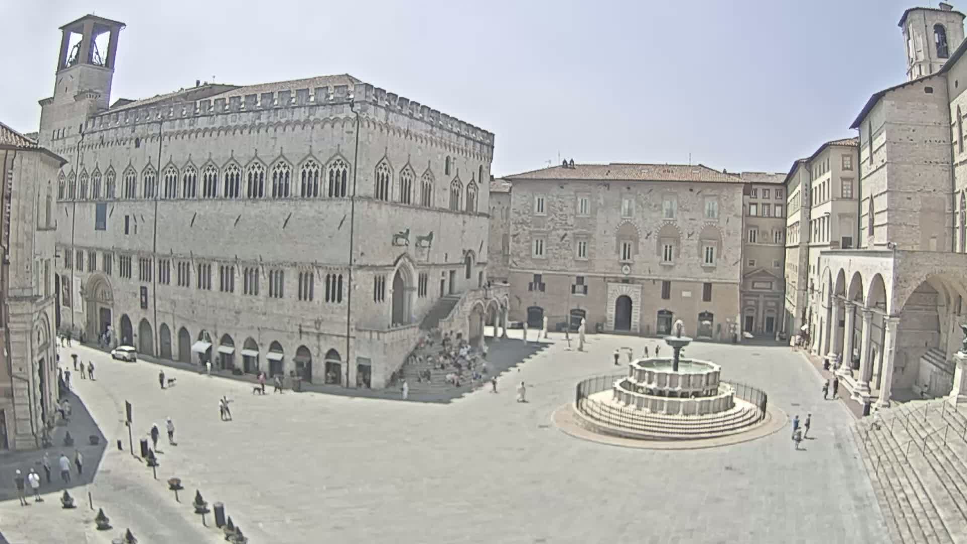 A large, sunny town square features a central fountain, surrounded by several stone buildings, one of which is a large palace with a tower.