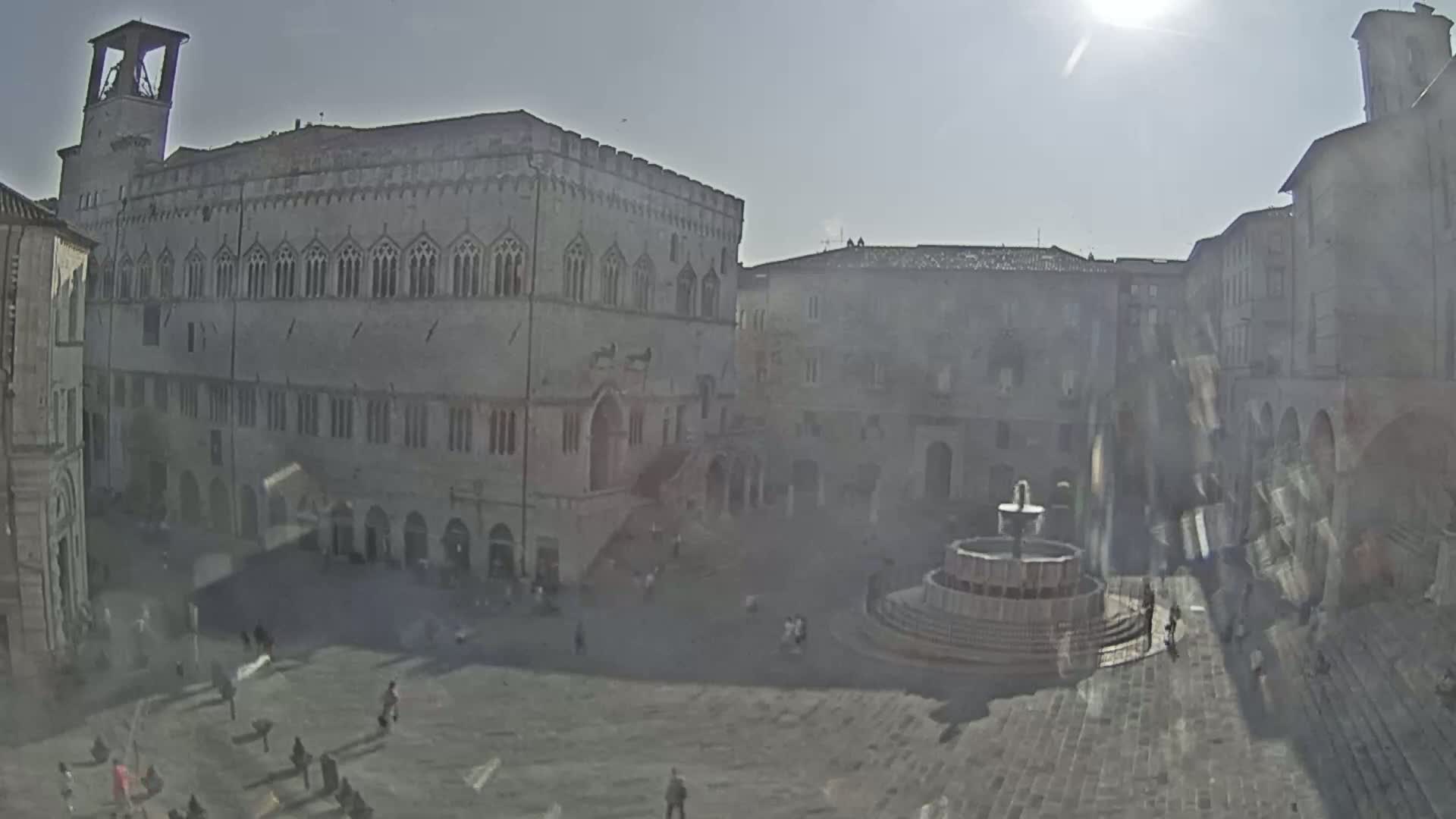 A sunny day reveals a large town square with a central fountain, surrounded by several multi-story stone buildings.