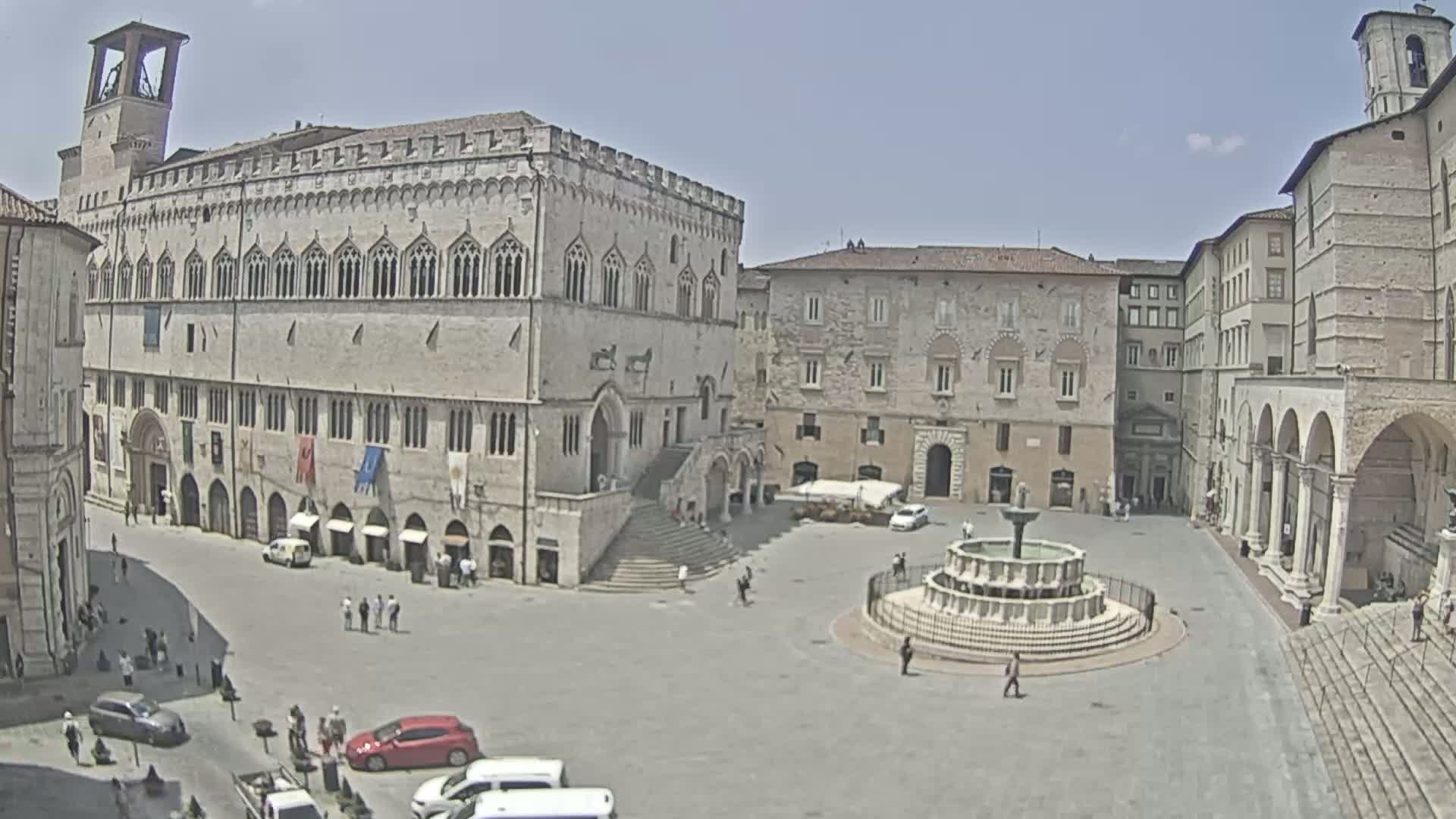 A large, sunlit town square contains a fountain, several historic stone buildings, and a scattering of people and cars.
